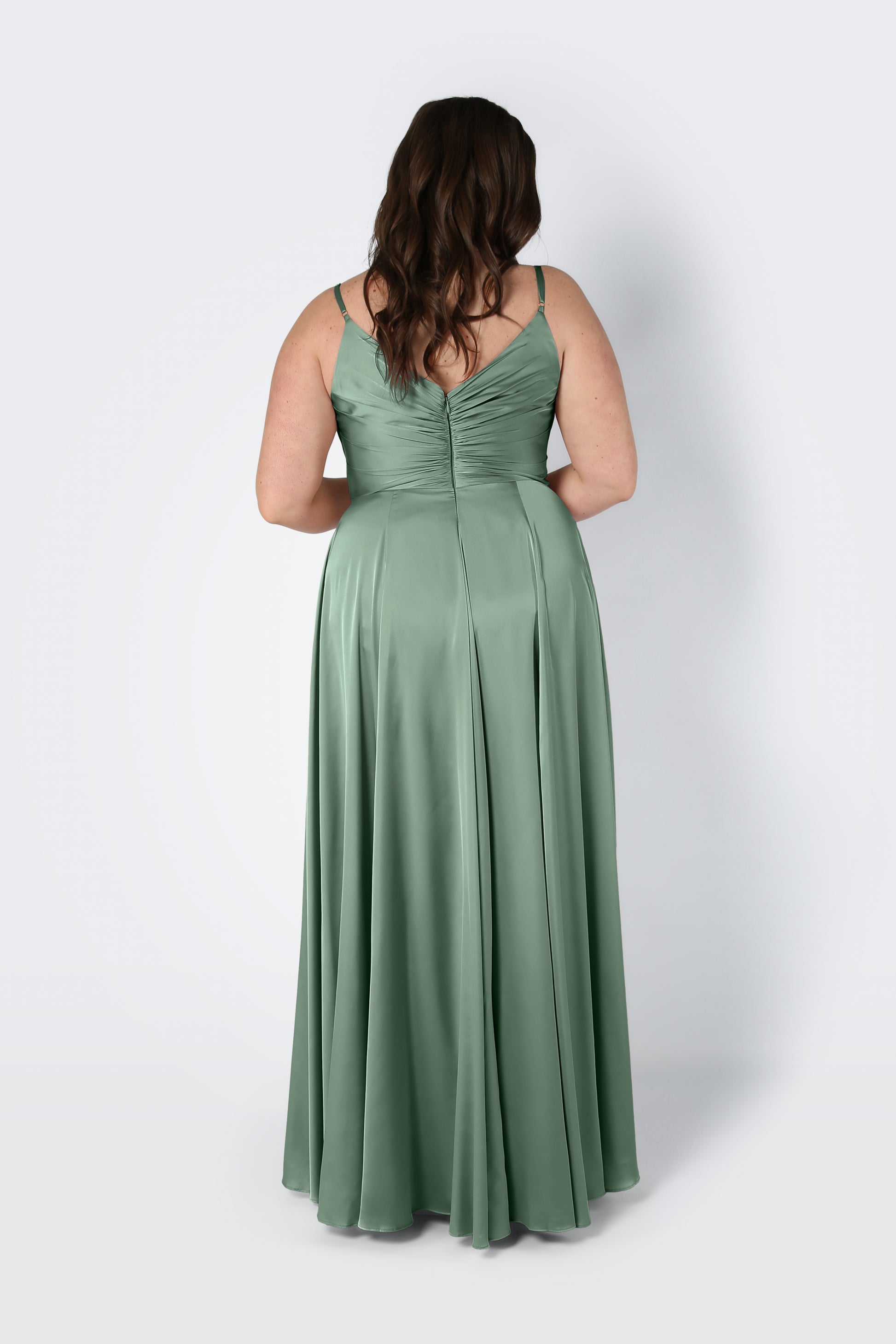 A woman with wavy brown hair, seen from behind, wears the TH&TH Isla Bridesmaid Dress in sage green satin—a long dress with thin straps and a fitted pleated bodice—standing against a plain light background.
