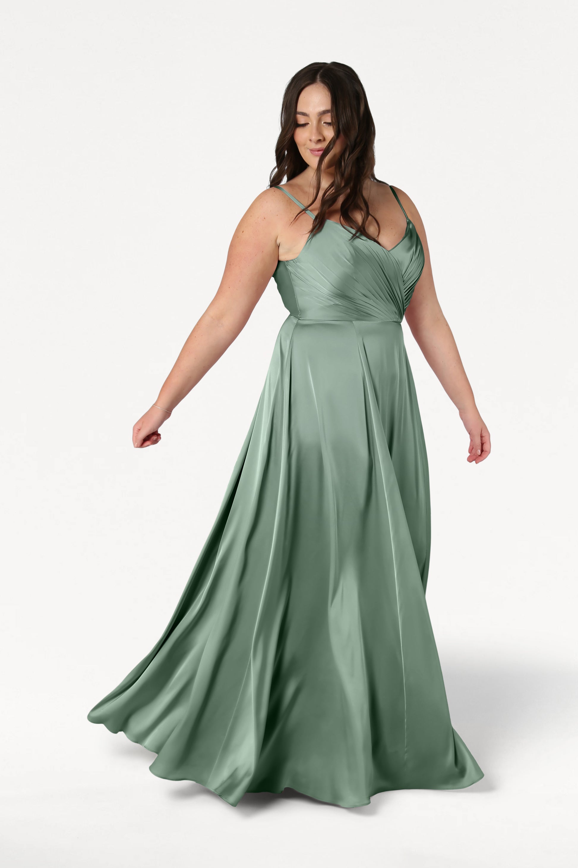 A woman twirls gracefully in the TH&TH Isla Bridesmaid Dress in Sage Green Satin, featuring spaghetti straps and a fitted, pleated bodice, set against a plain white background.