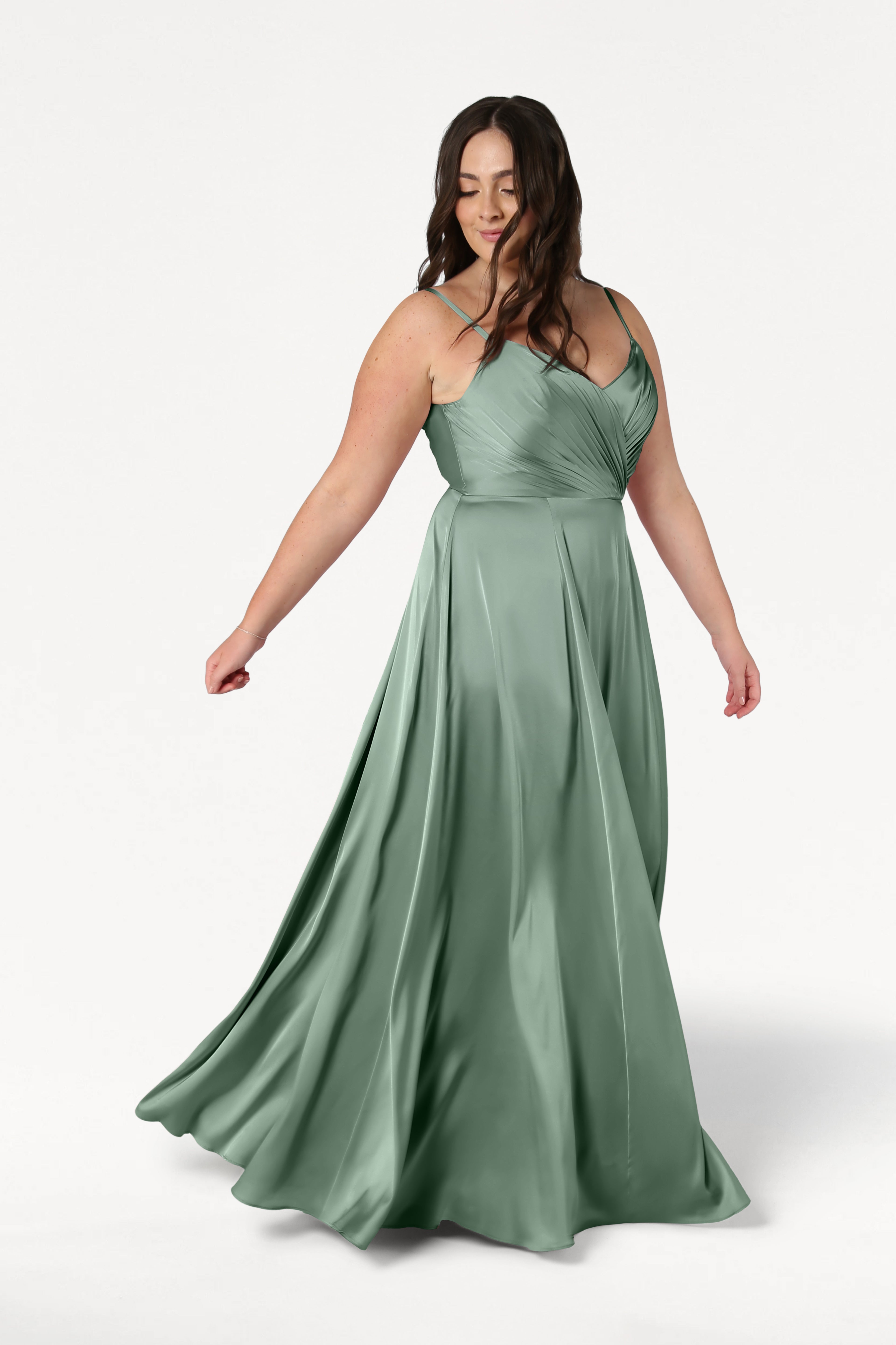 A woman twirls gracefully in the TH&TH Isla Bridesmaid Dress in Sage Green Satin, featuring spaghetti straps and a fitted, pleated bodice, set against a plain white background.