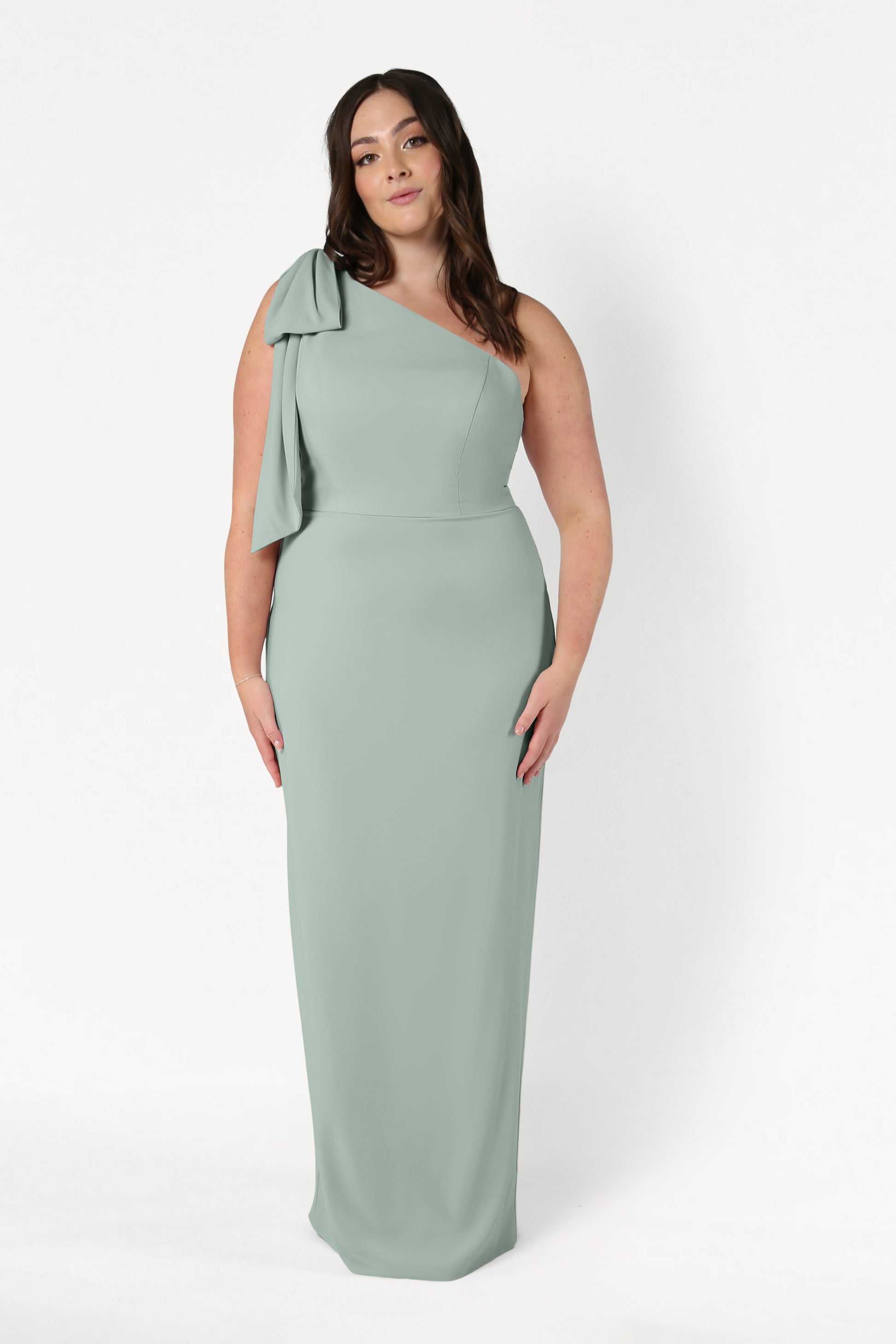 A woman with long brown hair stands against a white background in the TH&TH Valentina Bridesmaid Dress in Sage Green, a floor-length, one-shoulder crepe gown featuring a bow detail at the shoulder and a modern, elegant look.