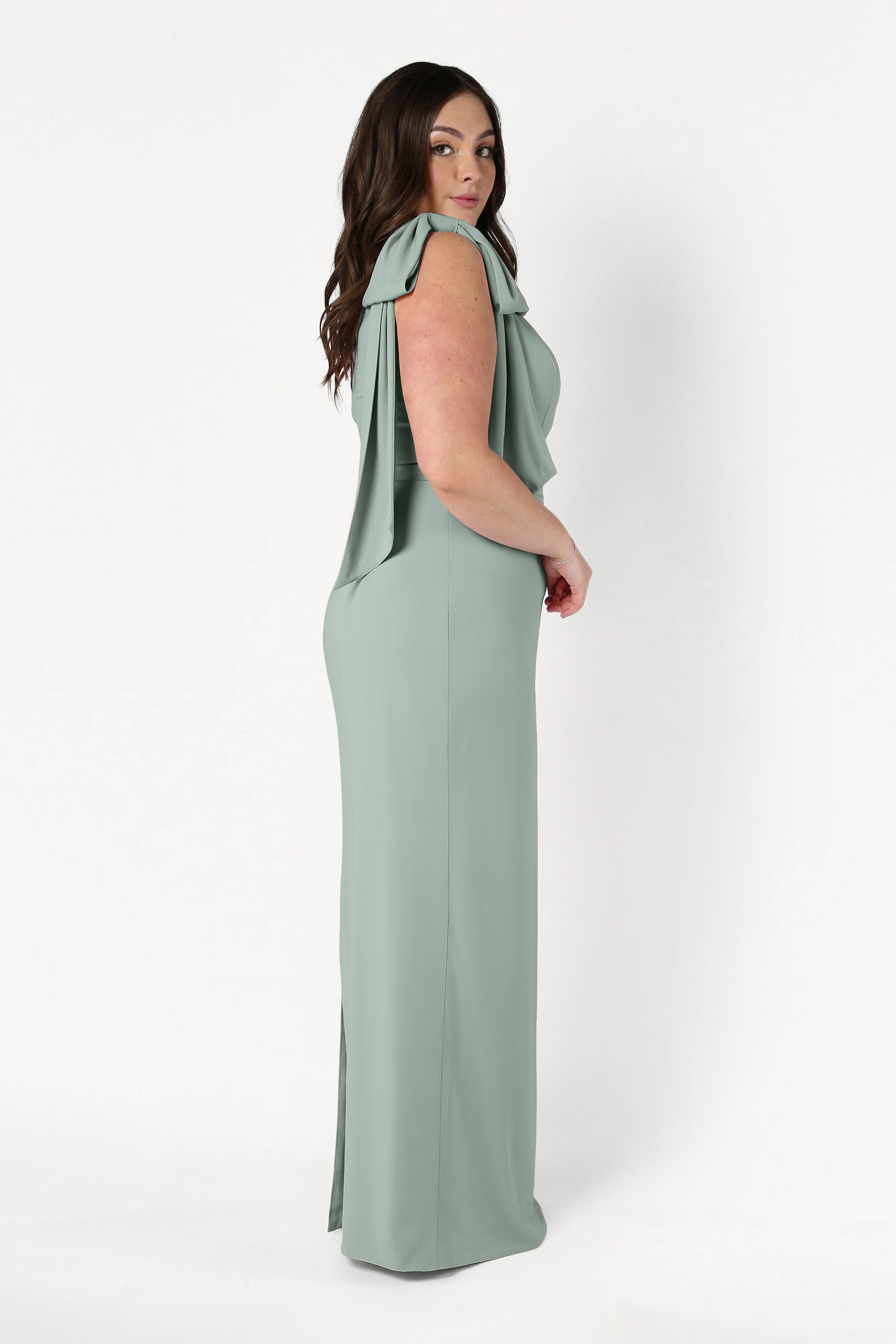 A woman with long dark hair stands in profile against a white background, wearing the TH&TH Valentina Bridesmaid Dress in Sage Green—a modern, size-inclusive crepe style with draped sleeves.