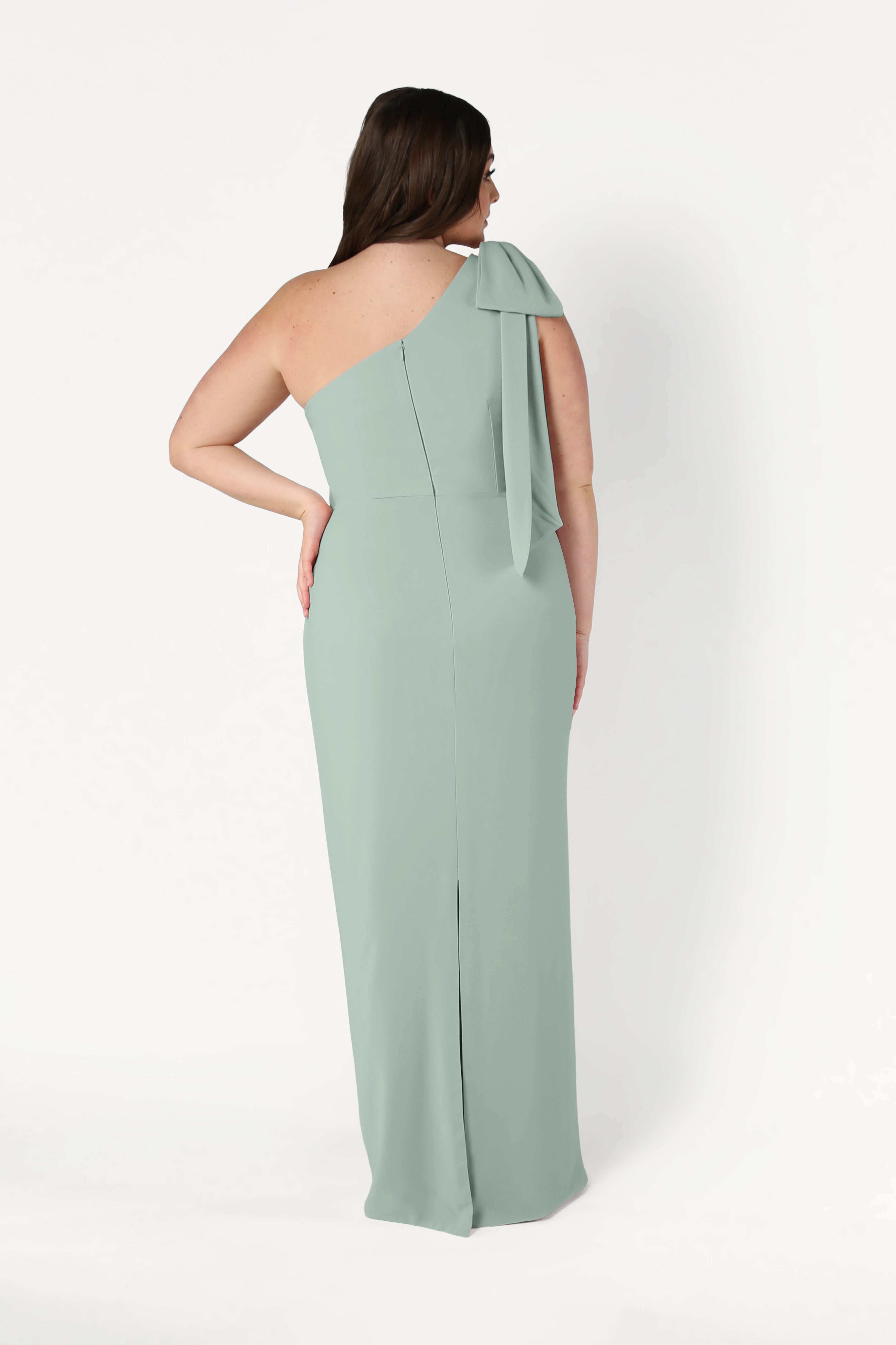 A woman stands with her back to the camera against a white background, wearing the TH&TH Valentina Bridesmaid Dress in sage green, featuring a one-shoulder design and bow detail.