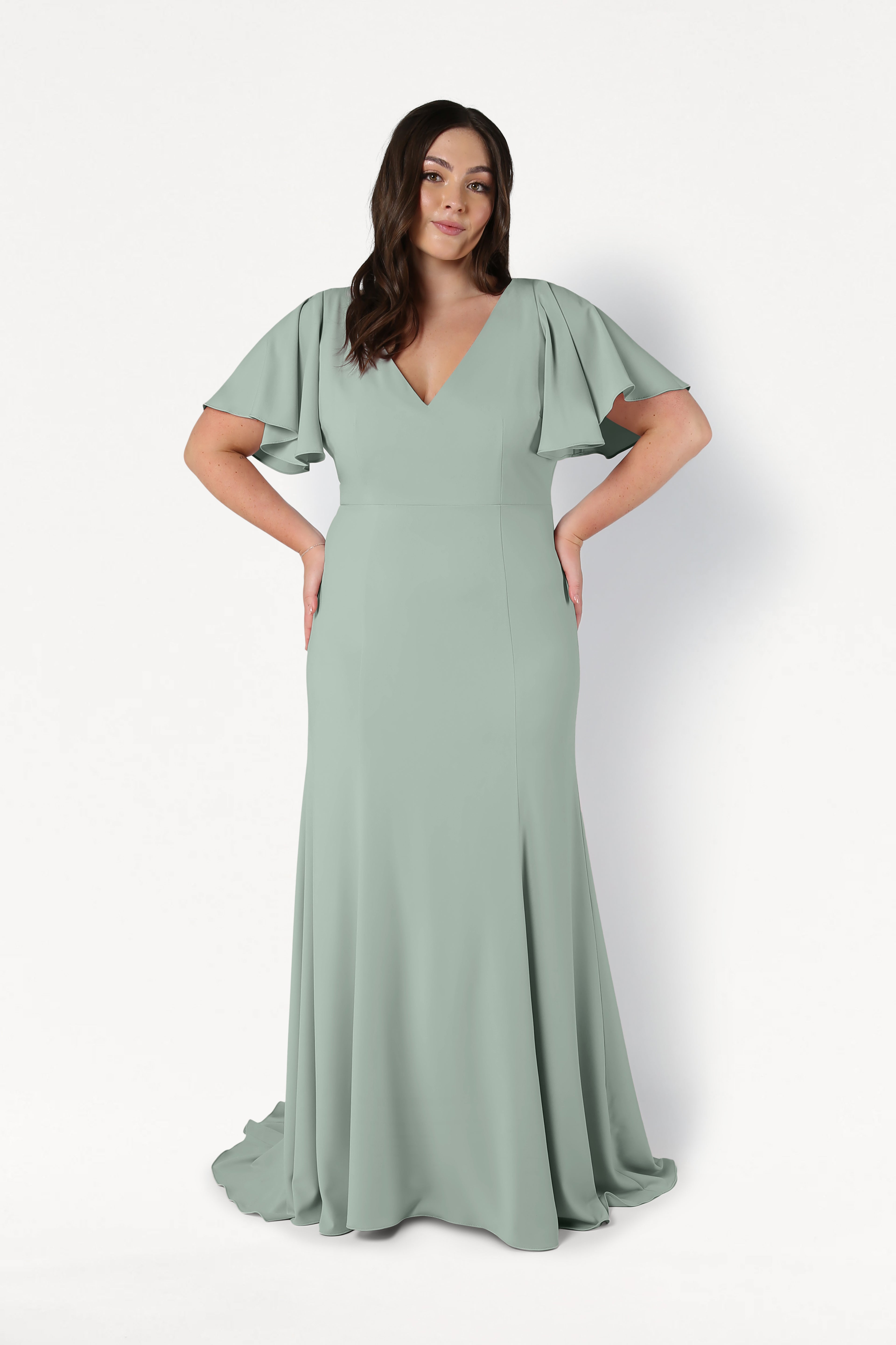 A woman with long, wavy dark hair smiles with her hands on her hips, wearing the TH&TH Celeste Moss Crepe Bridesmaid Dress in Sage Green—a floor-length gown with flutter sleeves and a V-neckline, standing against a white background.