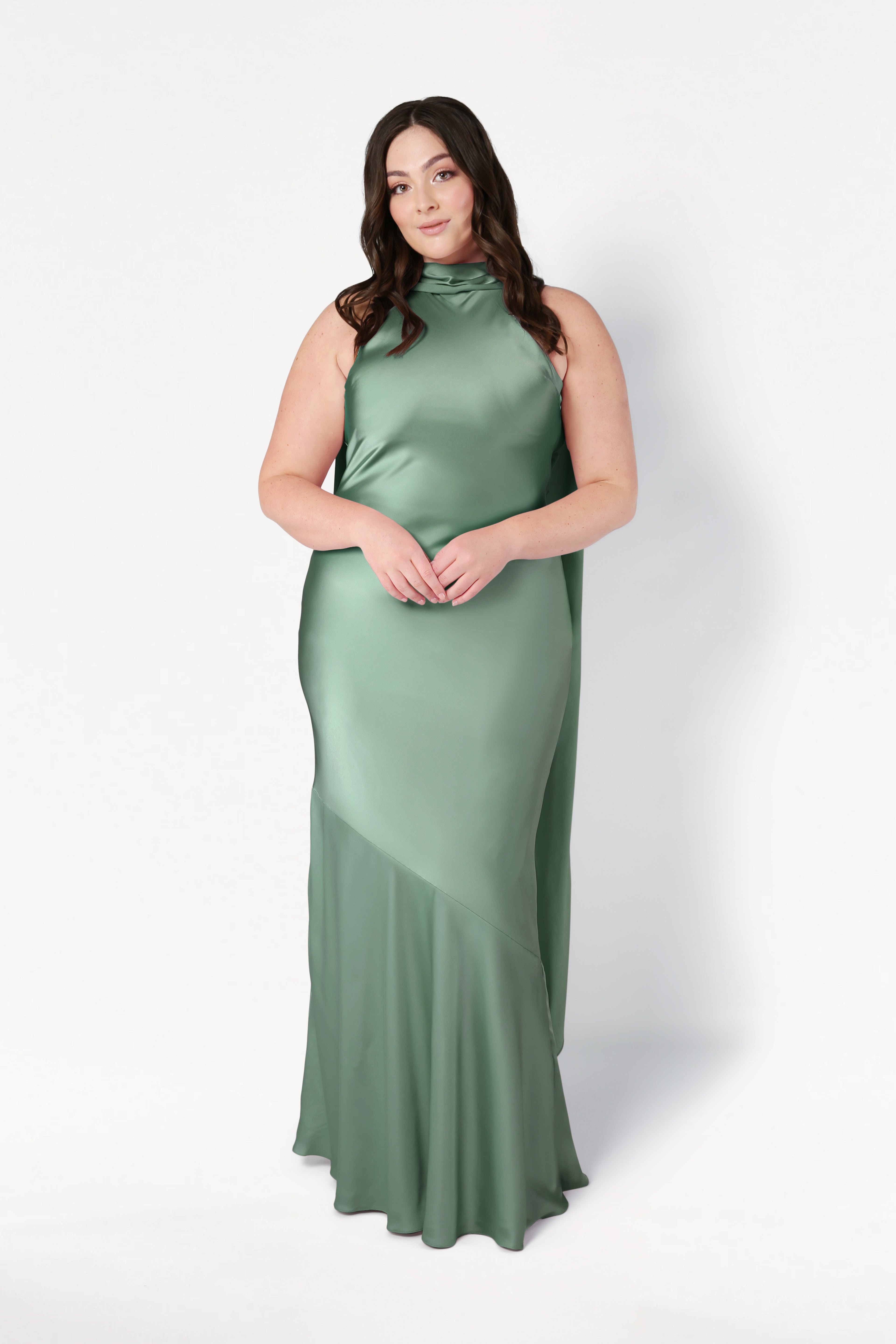 A woman with long dark hair wears the TH&TH Mira Bridesmaid Dress in sage green satin, a sleeveless, floor-length gown with a high neckline, standing against a plain white background.