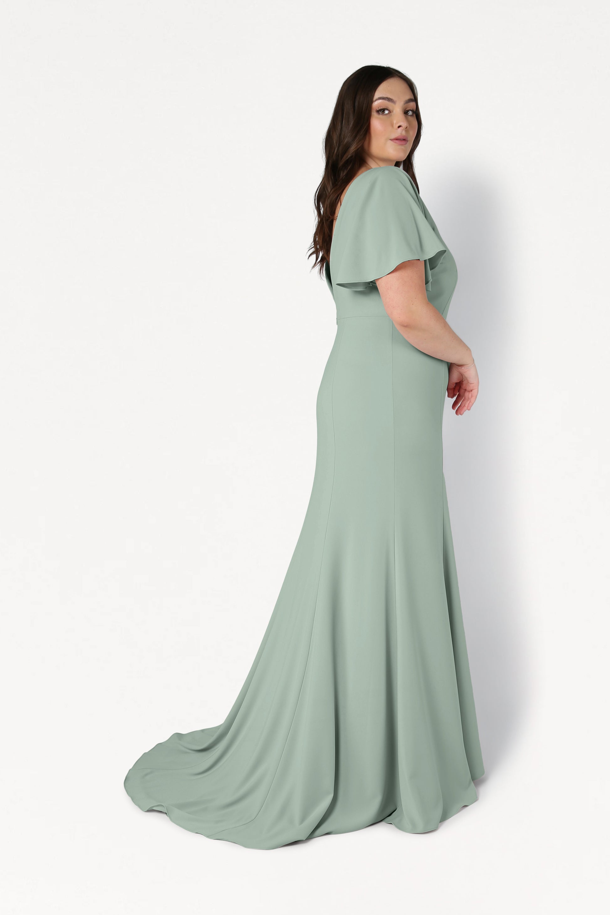 A woman stands in profile against a white background wearing the TH&TH Celeste Moss Crepe Bridesmaid Dress in Sage Green, featuring short flutter sleeves, a fitted bodice, and a flowing train.
