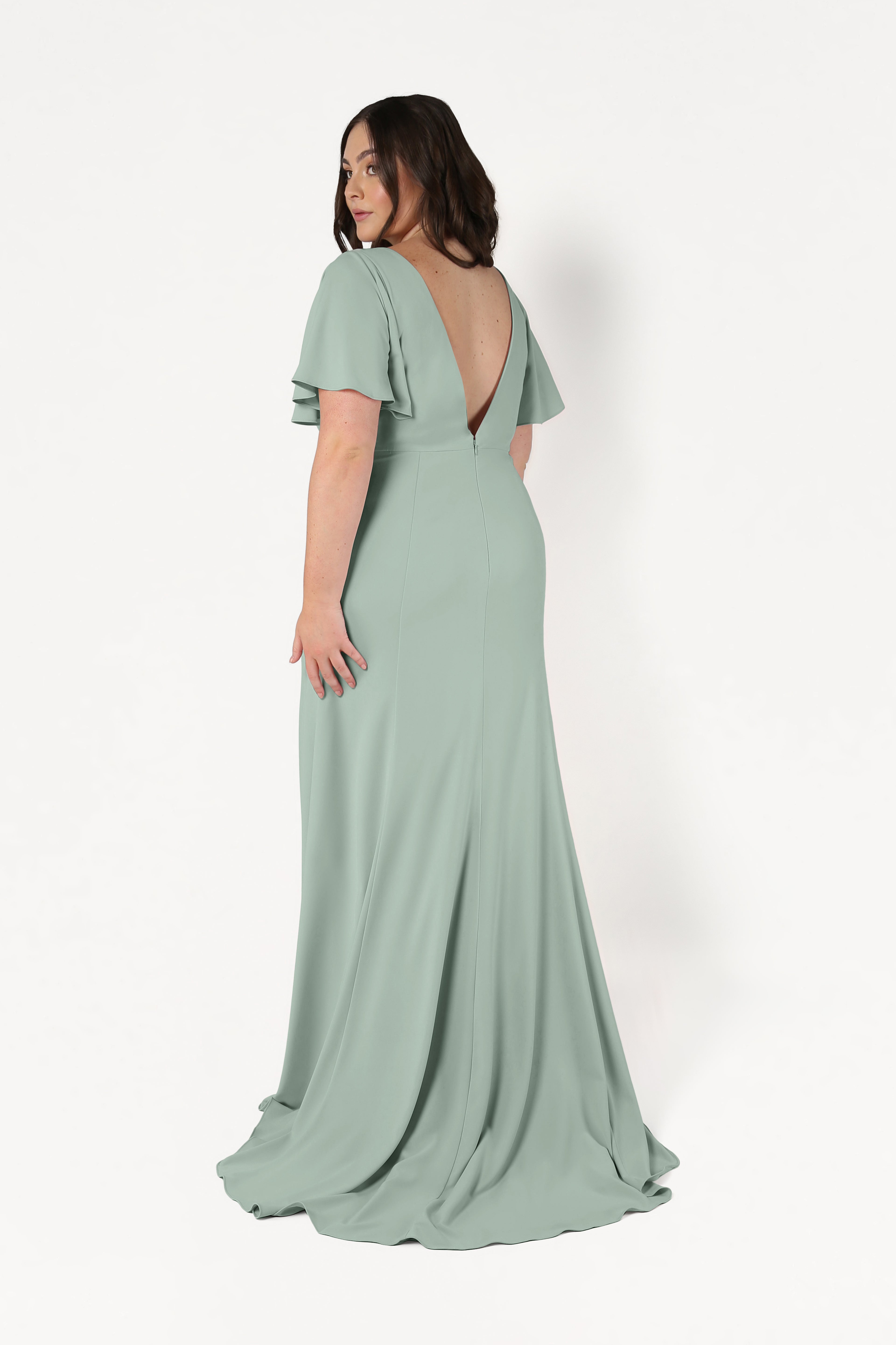 A woman models the TH&TH Celeste Moss Crepe Bridesmaid Dress in Sage Green, featuring flutter sleeves and a deep V-back, as she poses against a plain white background looking over her shoulder.