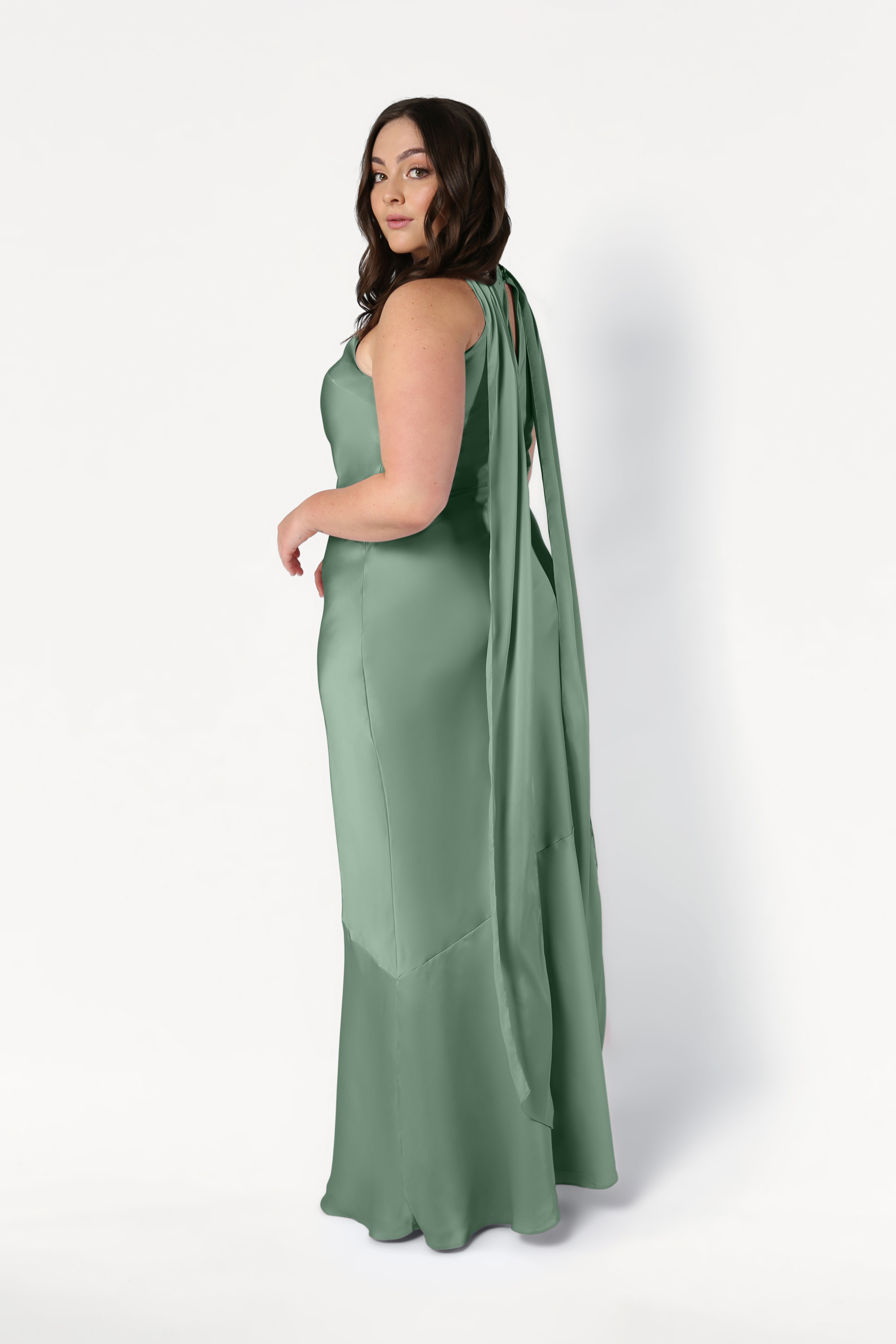 A woman with long brown hair models the TH&TH Mira Bridesmaid Dress in Sage Green Satin, a sleeveless, floor-length, bias-cut design with an elegant draped back. She stands sideways, looking over her shoulder against a plain white background.