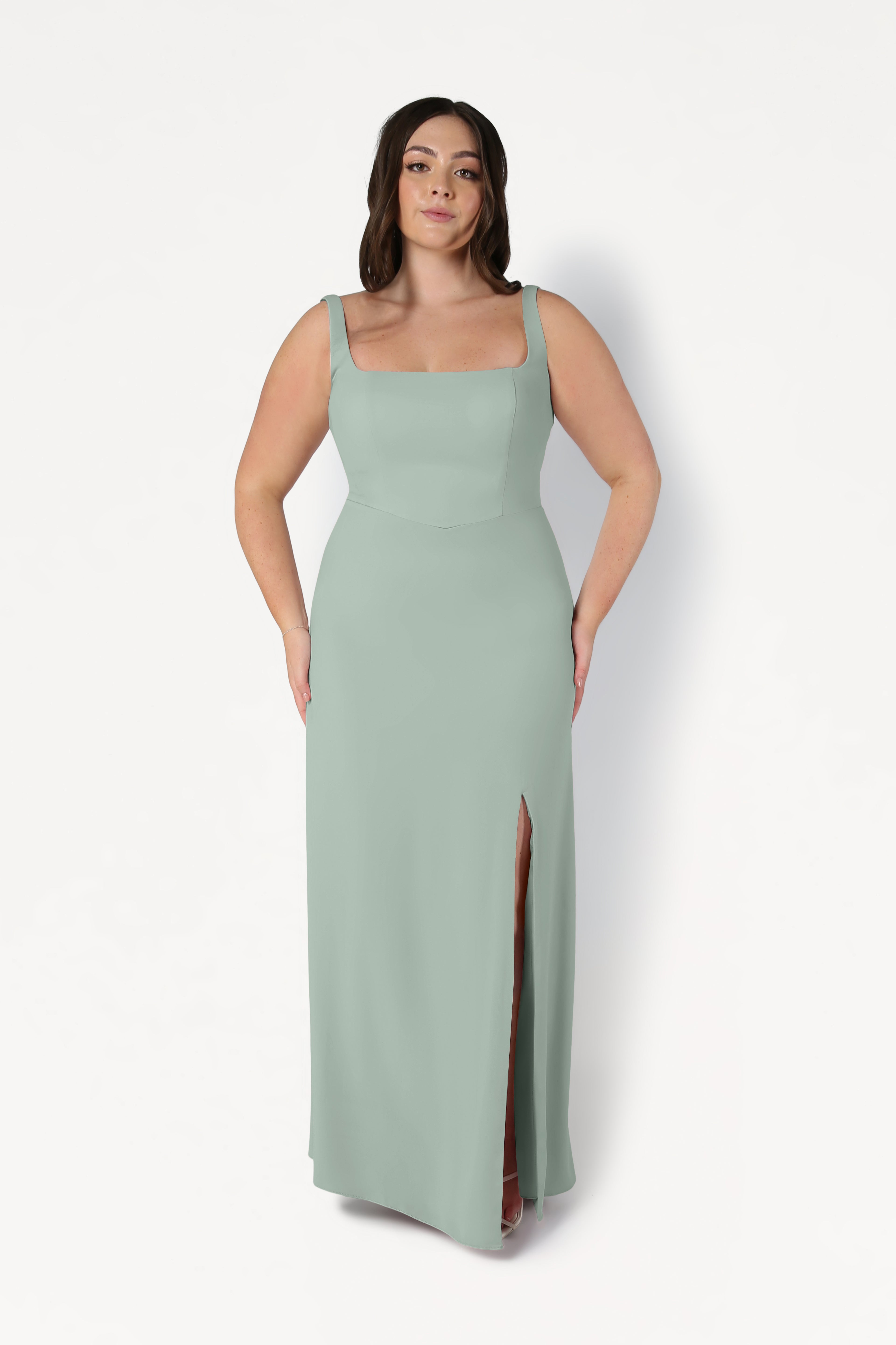 A woman models the TH&TH Helena Bridesmaid Dress in sage green, featuring a sleeveless, floor-length design with a square neckline and thigh-high slit, standing against a plain white background.