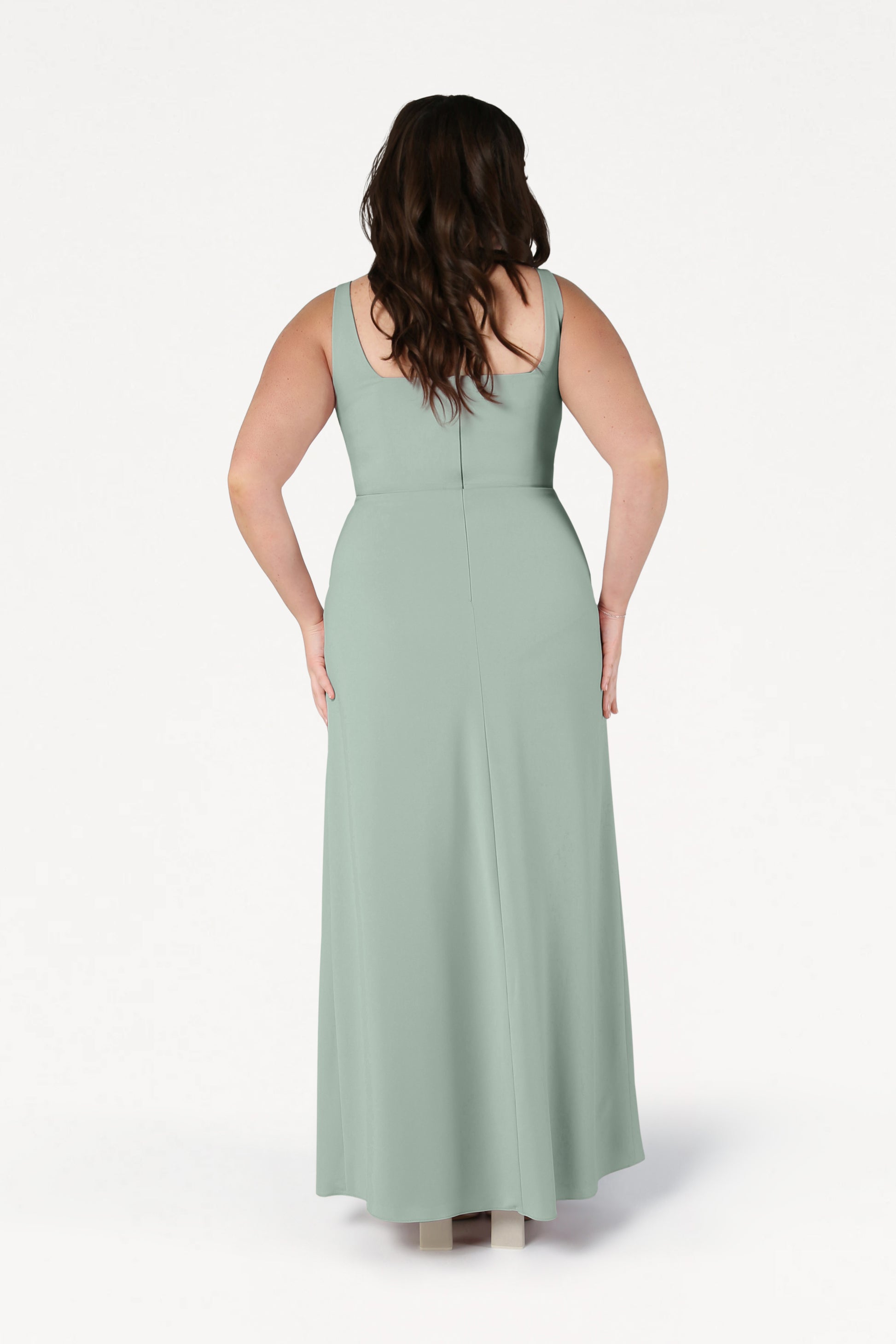A woman with long brown hair stands facing away, wearing the TH&TH Helena Bridesmaid Dress in Sage Green—a modern, sleeveless, floor-length gown made from moss crepe. The background is plain white.