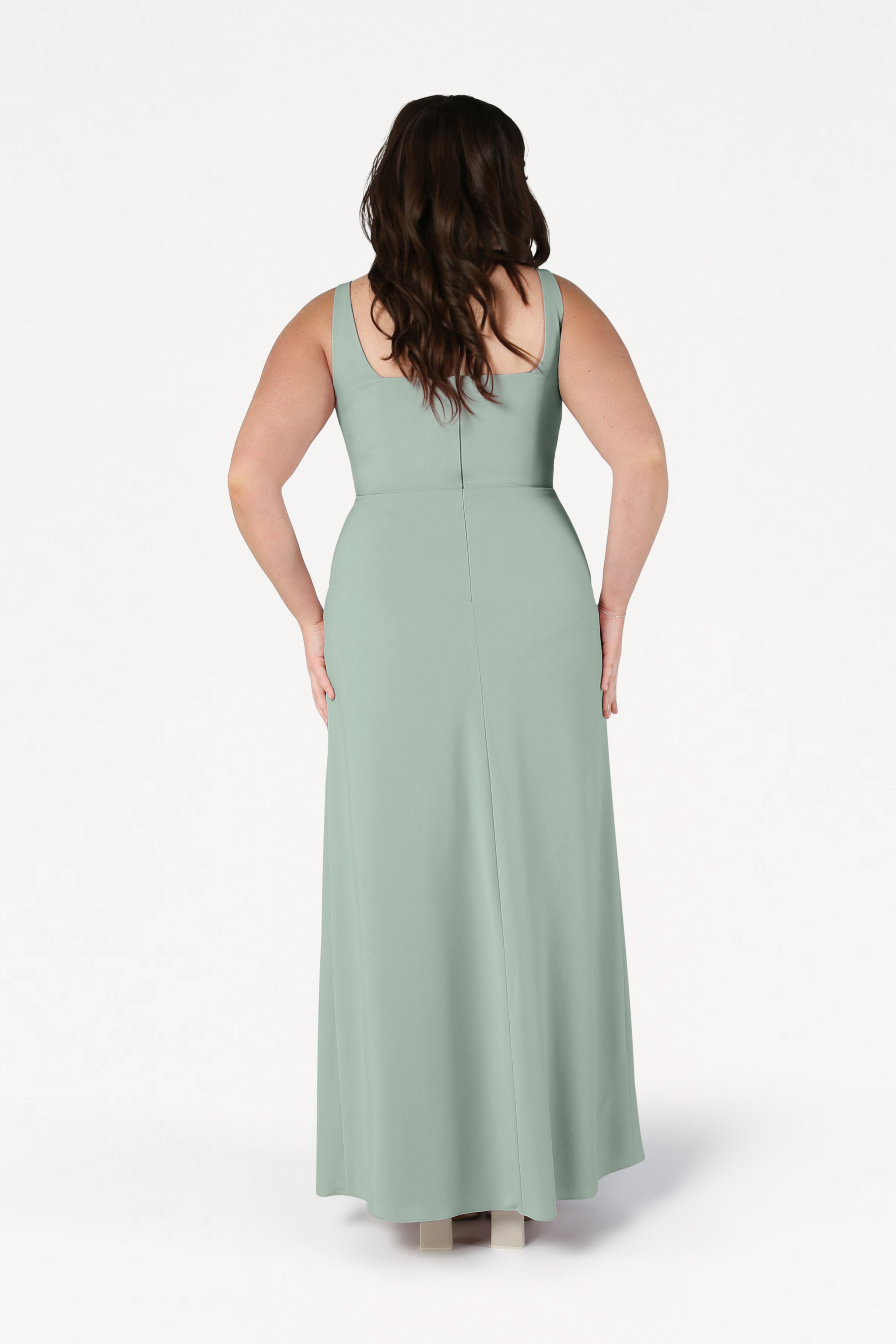 A woman with long brown hair stands facing away, wearing the TH&TH Helena Bridesmaid Dress in Sage Green—a modern, sleeveless, floor-length gown made from moss crepe. The background is plain white.