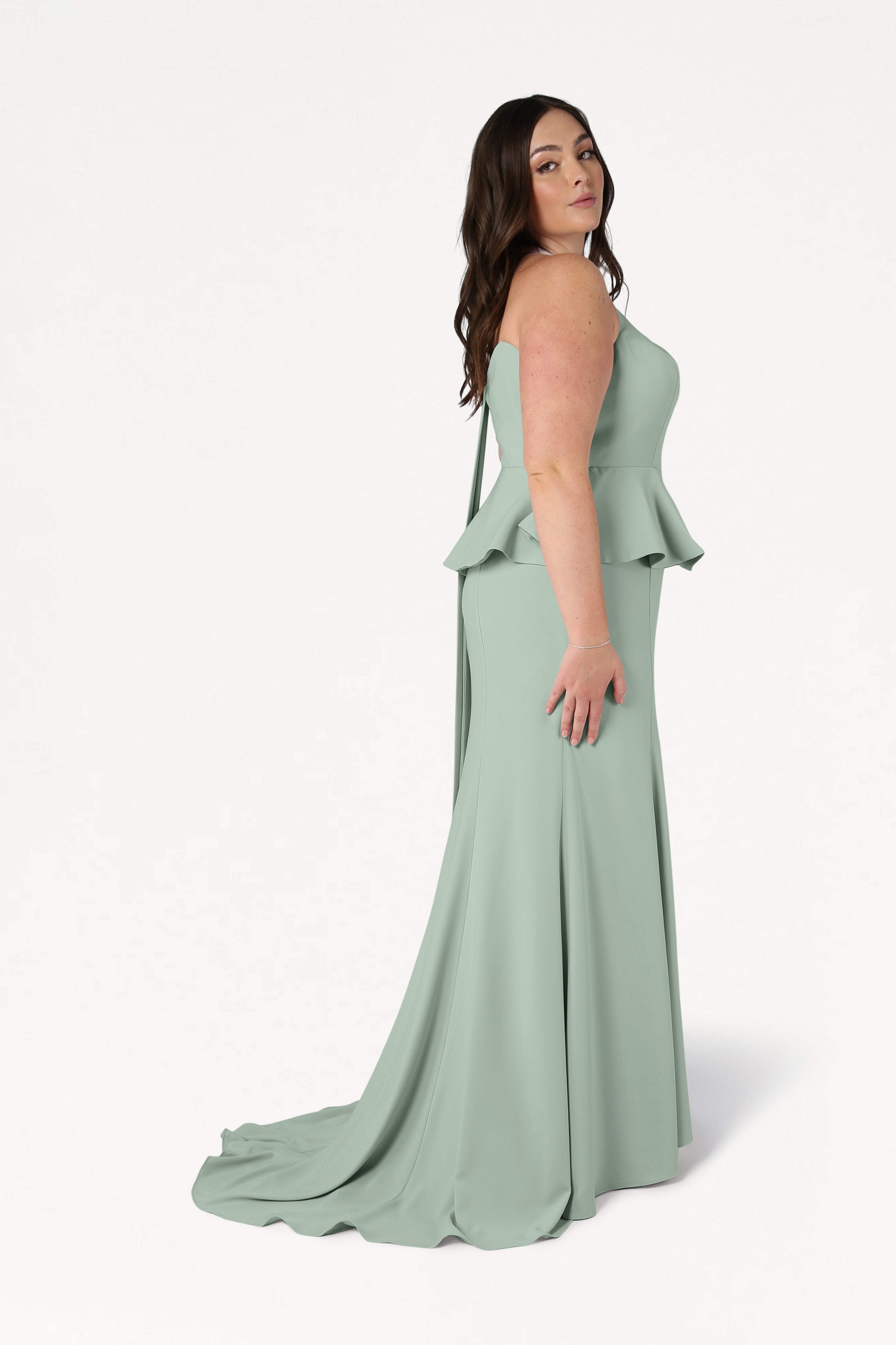 A woman with long brown hair stands sideways against a plain white background, wearing the TH&TH Peony Bridesmaid Dress in Sage Green—a modern strapless moss crepe gown with a peplum and floor-length skirt.