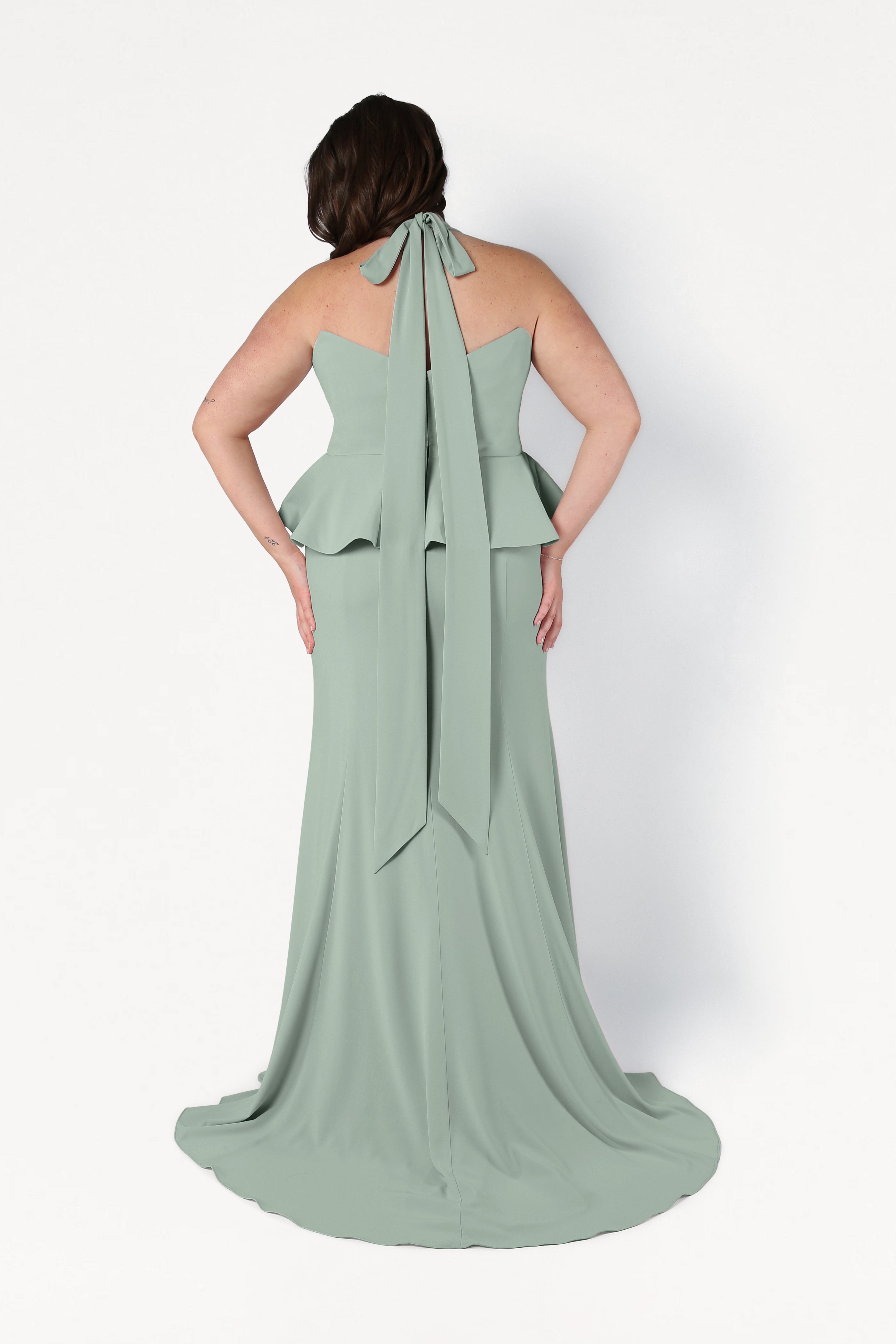A woman faces away, showcasing the back details of the TH&TH Peony Bridesmaid Dress in Sage Green, a floor-length gown with a peplum design and long neck tie, set against a plain white background.
