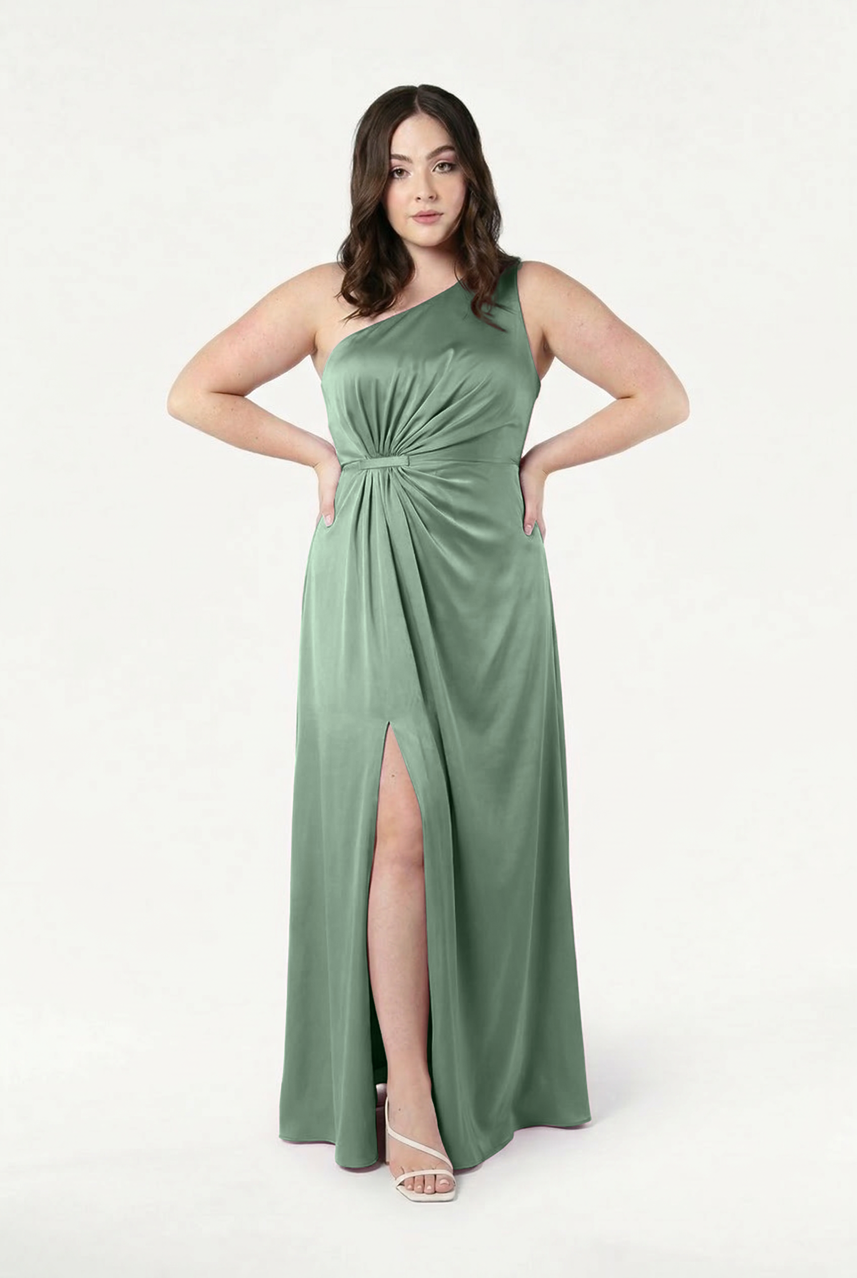 A woman wears the TH&TH Greta Bridesmaid Dress in Sage Green Satin, featuring a one-shoulder Grecian drape and waist knot. She poses with hands on hips, styled with wavy brown hair and white heeled sandals against a plain white background.