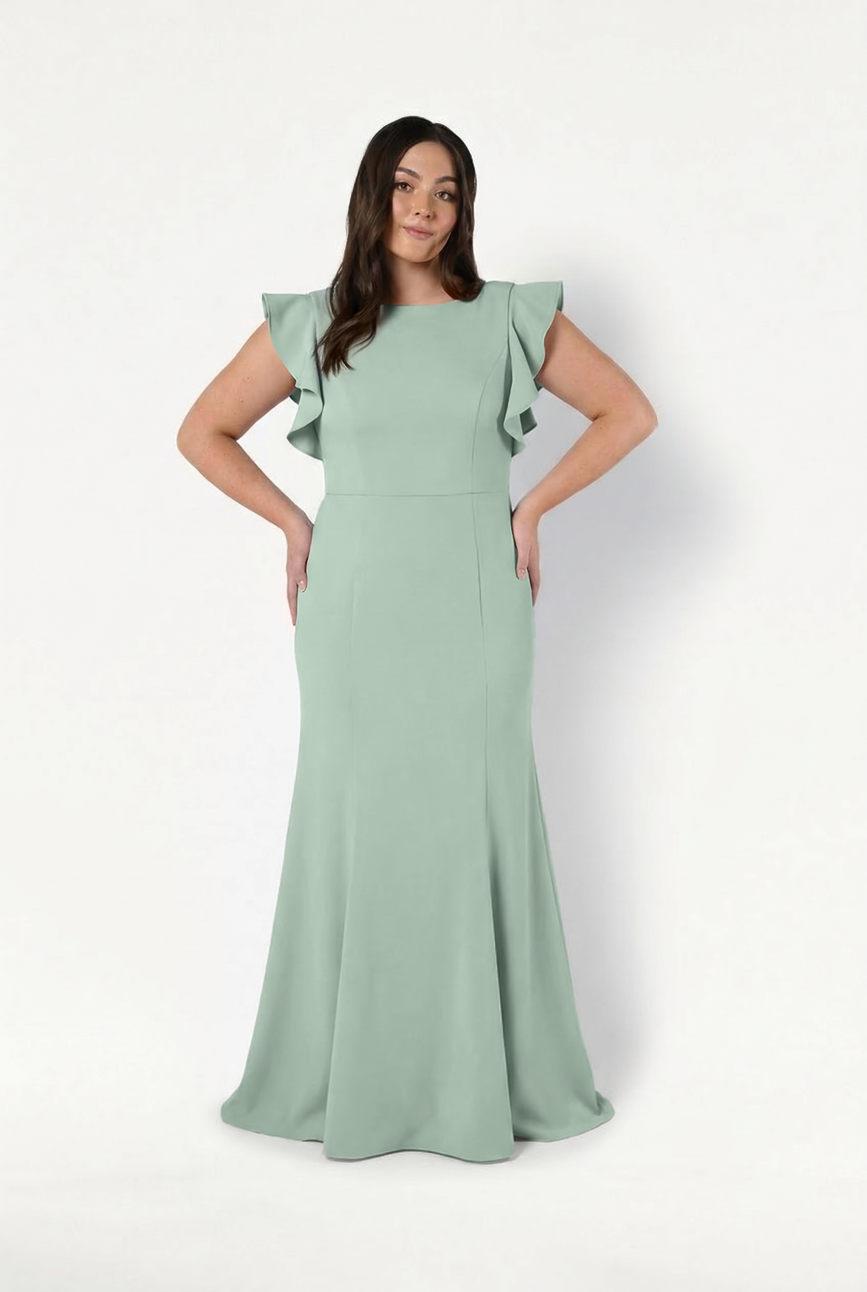 A woman poses against a white background in the floor-length Cecelia Moss Crepe Bridesmaid Dress in Sage Green by TH&TH, featuring ruffled short sleeves and a fitted bodice. She smiles softly with her hands on her hips.