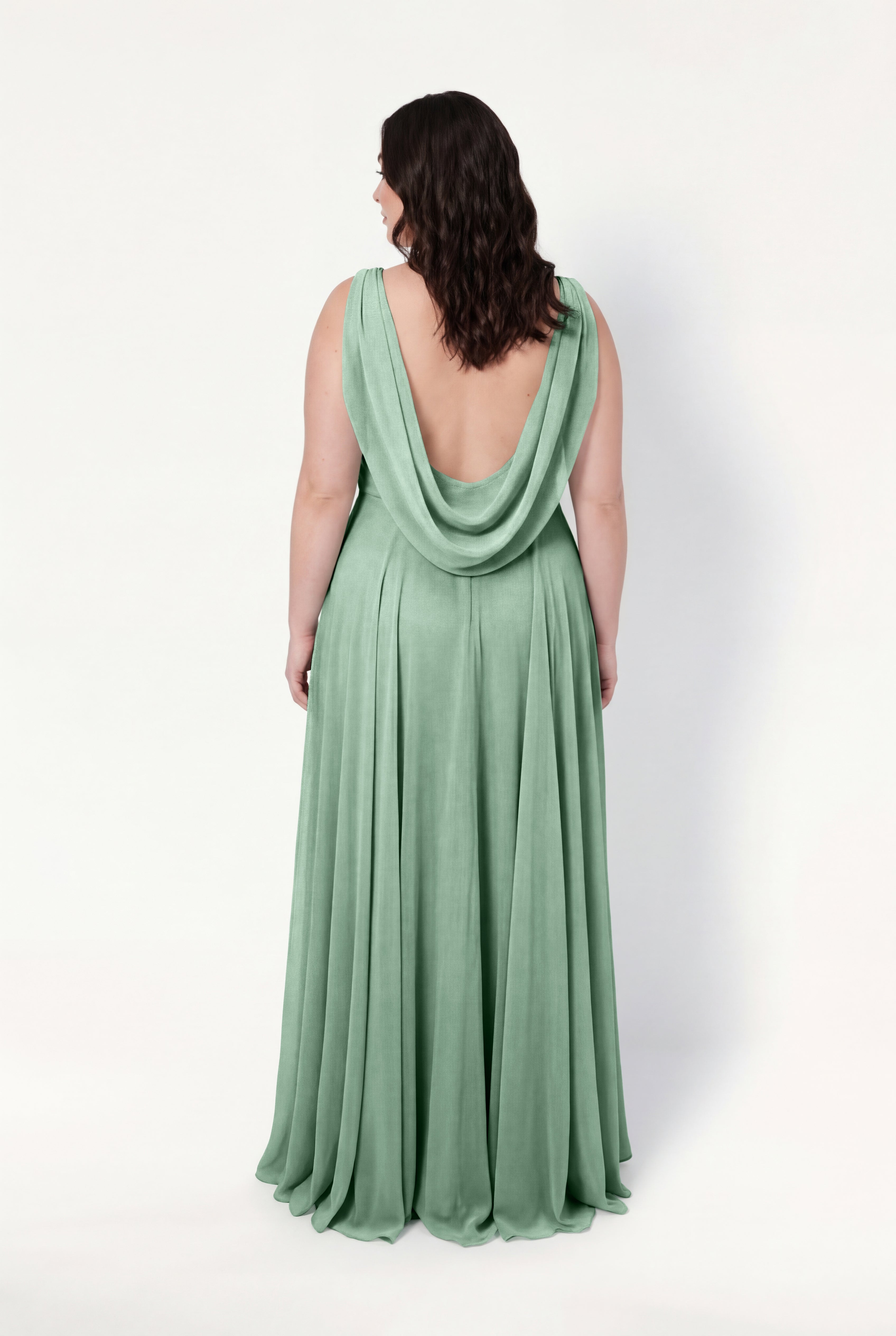 A woman with wavy brown hair faces away in the TH&TH Athena Bridesmaid Dress, a long, sleeveless sage green chiffon gown with a deep draped back, set against a plain white background.