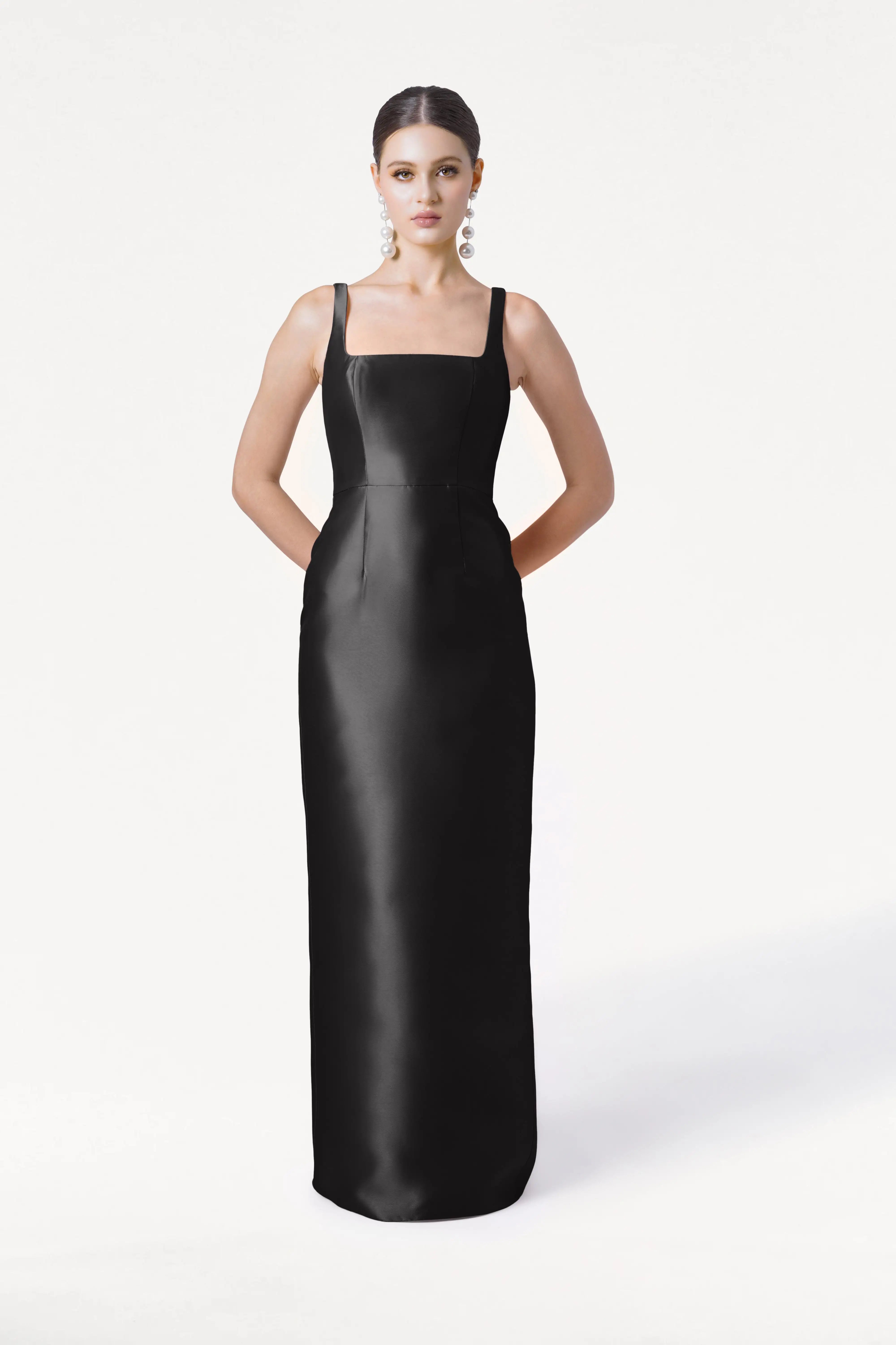 A woman poses against a white background, wearing the TH&TH Savoy Bridesmaid Dress in Black Satin Twill—a sleeveless, floor-length gown with a square neckline. She completes the look with large round earrings and neatly styled back hair.