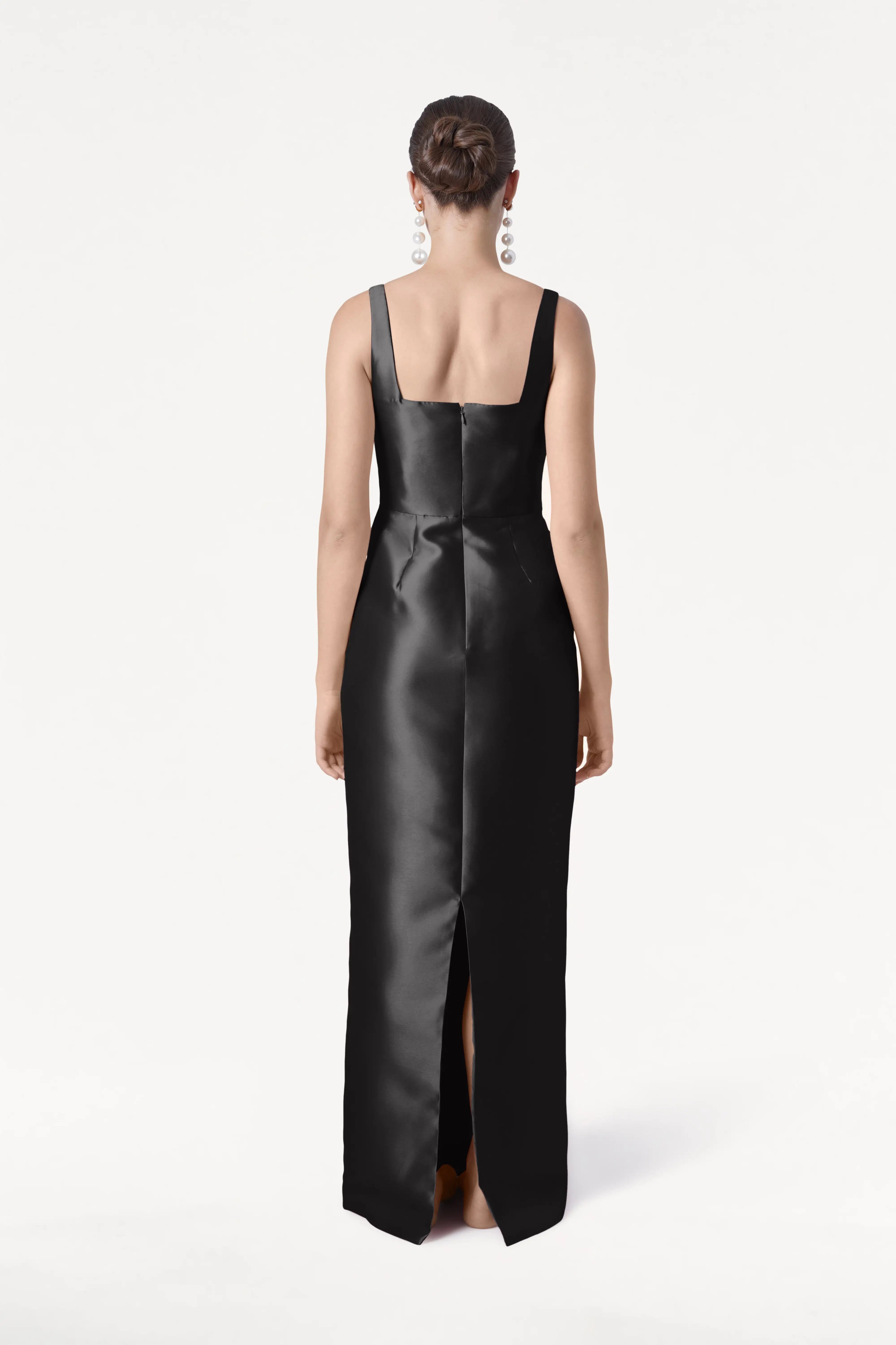 A woman faces away, wearing the TH&TH Savoy Bridesmaid Dress in Black Satin Twill. Her hair is in a neat bun, she has statement earrings, and the modern dress features a square neckline and back slit.