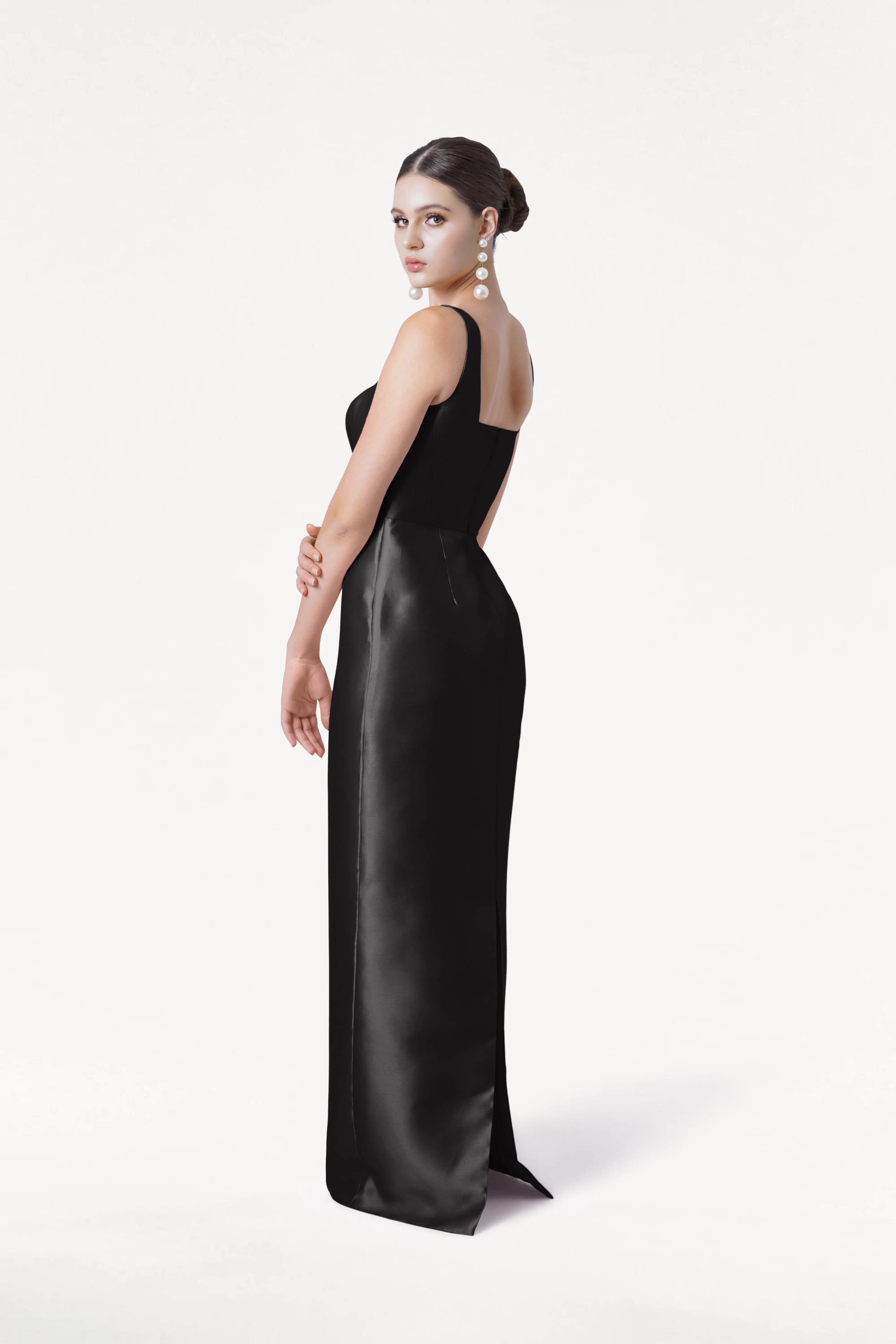 A woman with dark hair in an elegant bun models the TH&TH Savoy Bridesmaid Dress in Black Satin Twill and large pearl earrings, posed against a plain white background.