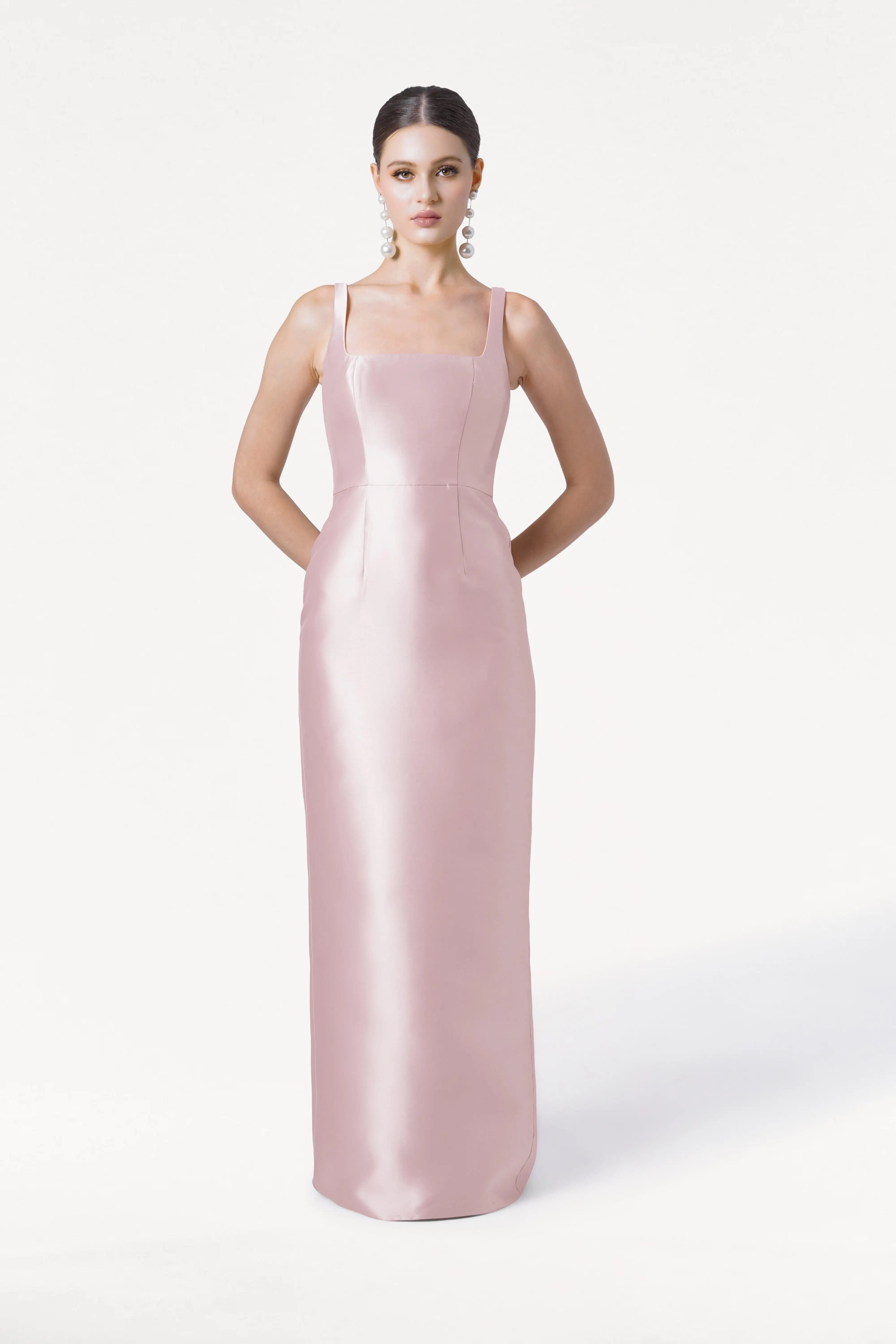 A woman poses against a white background in the TH&TH Savoy Bridesmaid Dress, crafted from blush pink satin twill with a modern square neckline and thin straps. Her hair is slicked back and she wears large statement earrings, with her hands behind her back.