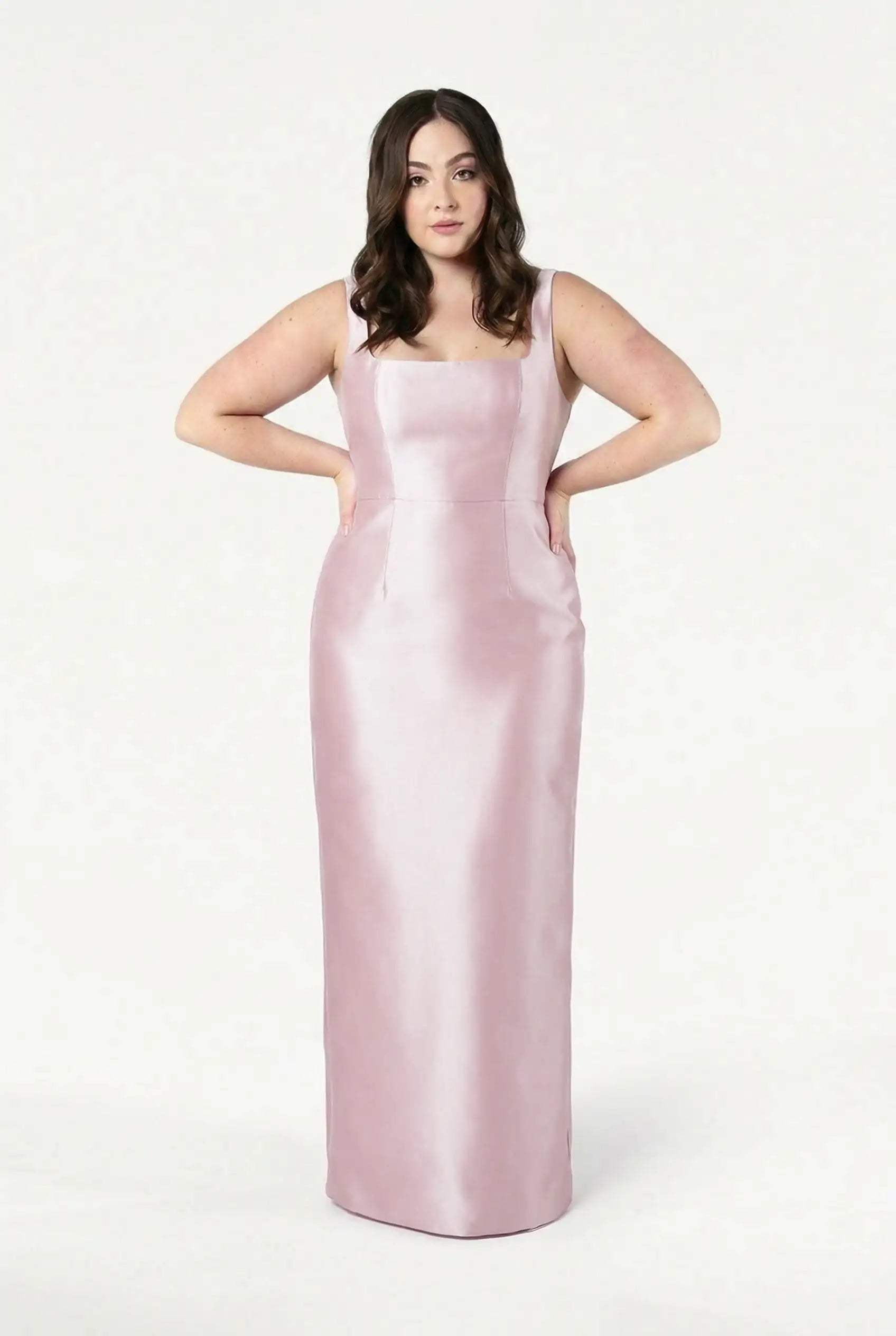 A woman with wavy brown hair stands facing forward against a white background, wearing the TH&TH Savoy Bridesmaid Dress in blush pink satin twill—a sleeveless, floor-length gown with a modern square neckline.