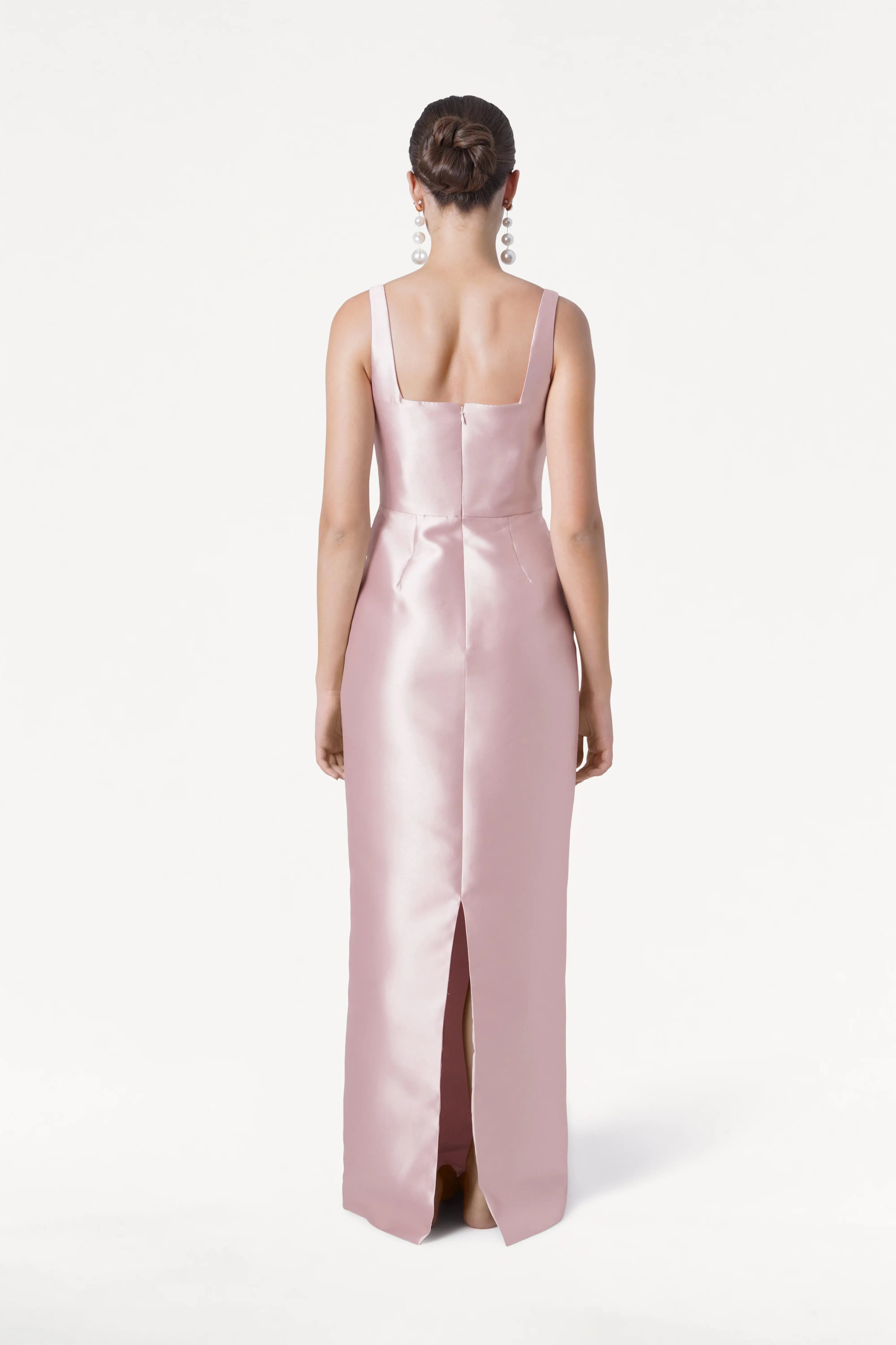 A woman models the TH&TH Savoy Bridesmaid Dress in Blush Pink Satin Twill, featuring a sleeveless silhouette, modern square neckline, and back slit, as she stands facing away against a plain white background.
