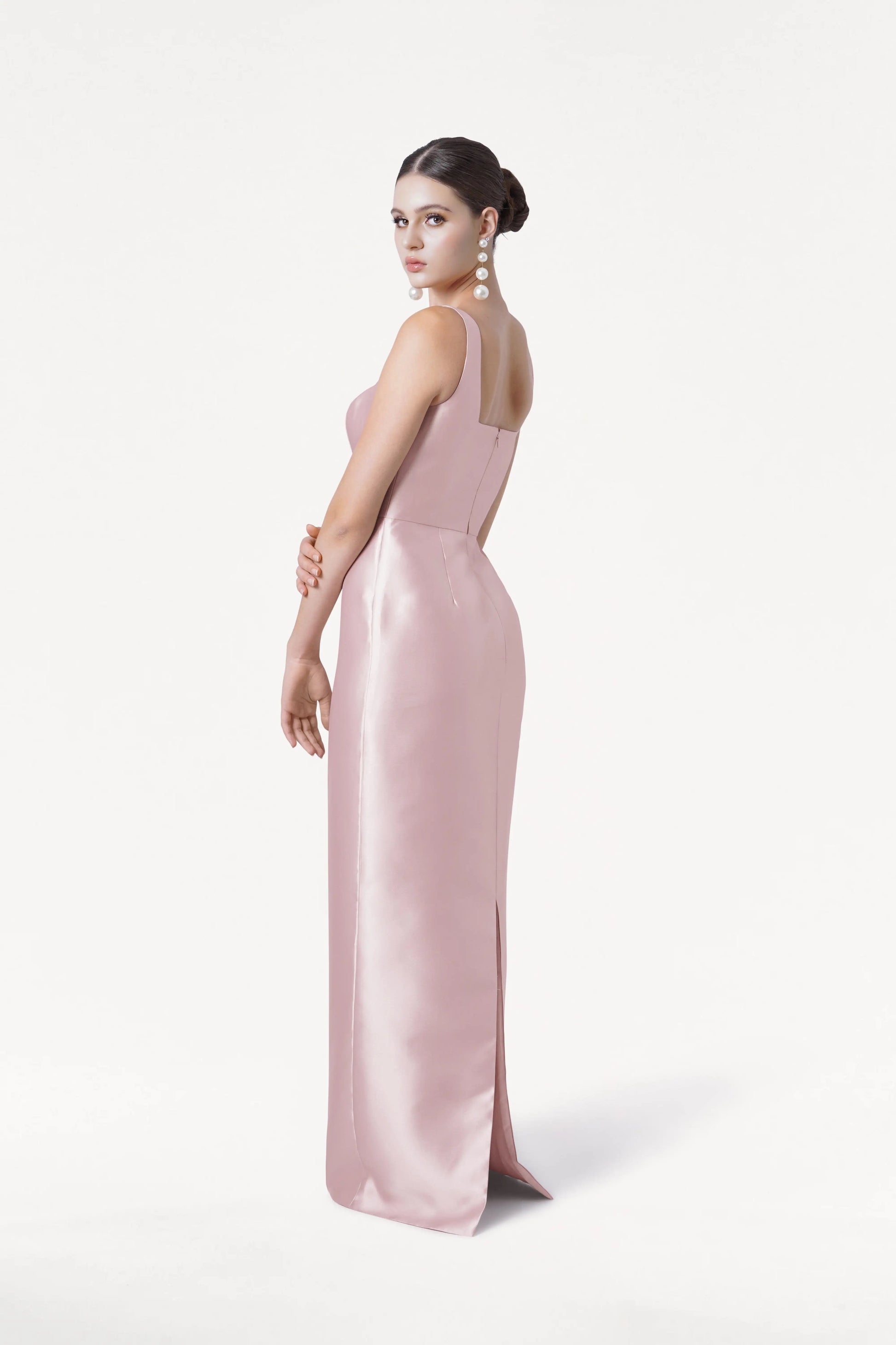 A woman models the TH&TH Savoy Bridesmaid Dress in blush pink satin twill, featuring a fitted bodice, long skirt, and modern square neckline. Her hair is up and she wears long earrings, posing sideways against a white background.