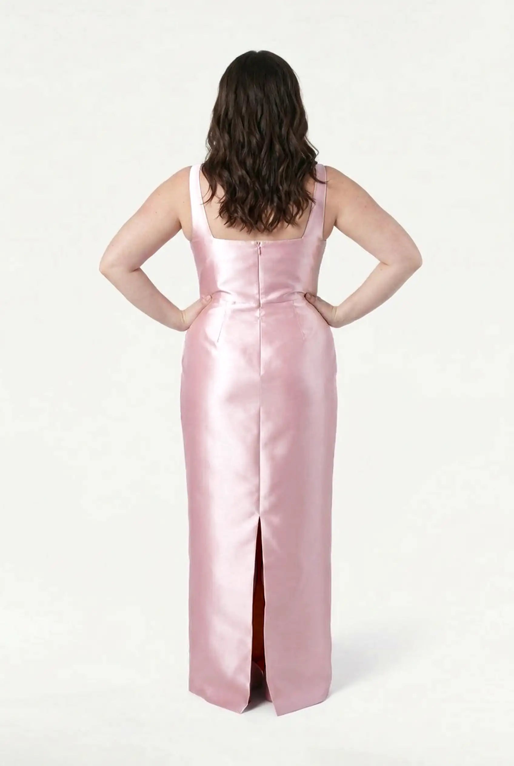 A person with wavy brown hair faces away, hands on hips, wearing the TH&TH Savoy Bridesmaid Dress in Blush Pink Satin Twill featuring a modern square neckline and back slit, set against a plain white background.
