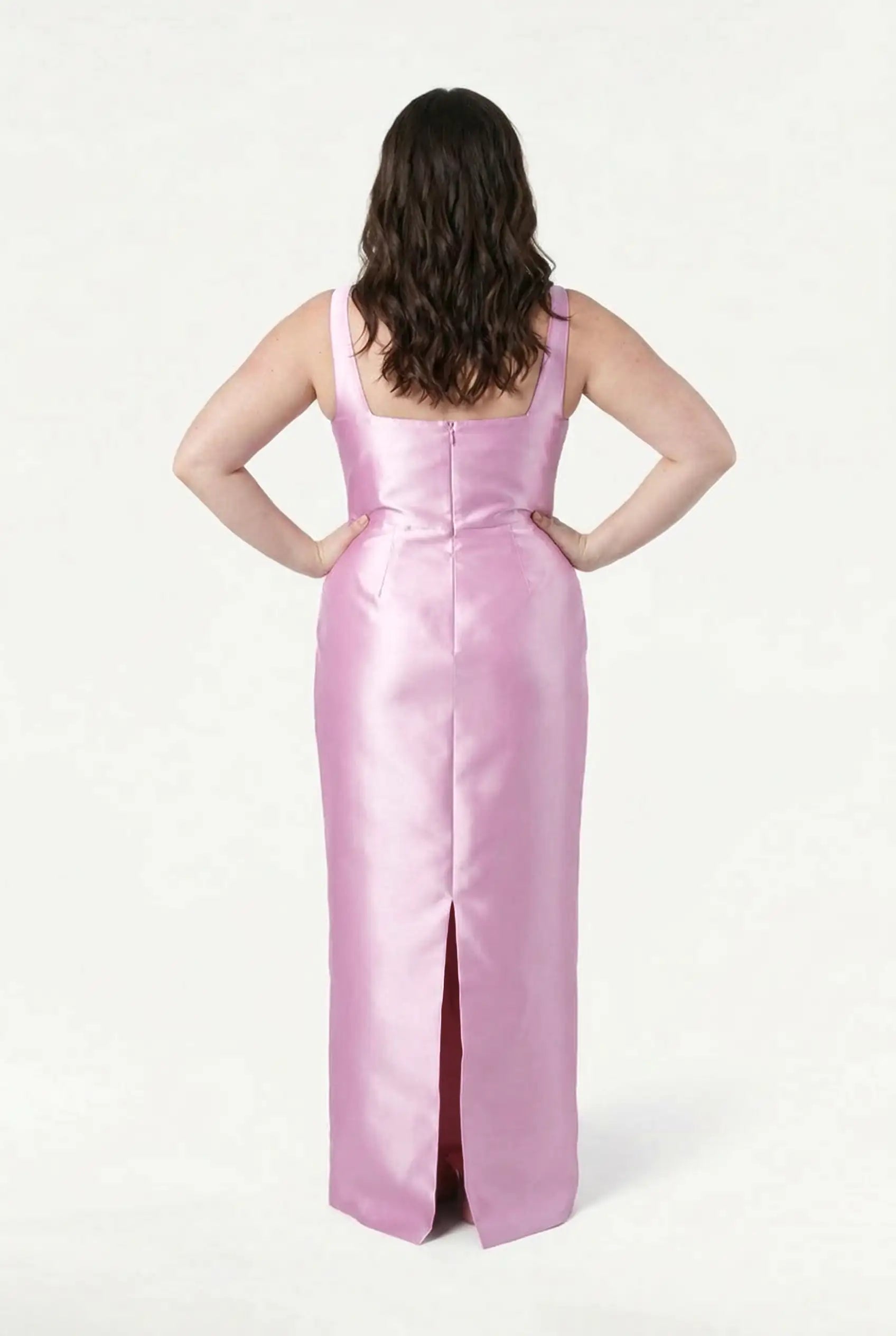 A woman with wavy brown hair stands hands on hips in the TH&TH Savoy Bridesmaid Dress, a sleeveless bubblegum pink satin twill gown with a back slit, photographed from behind against a plain white background.