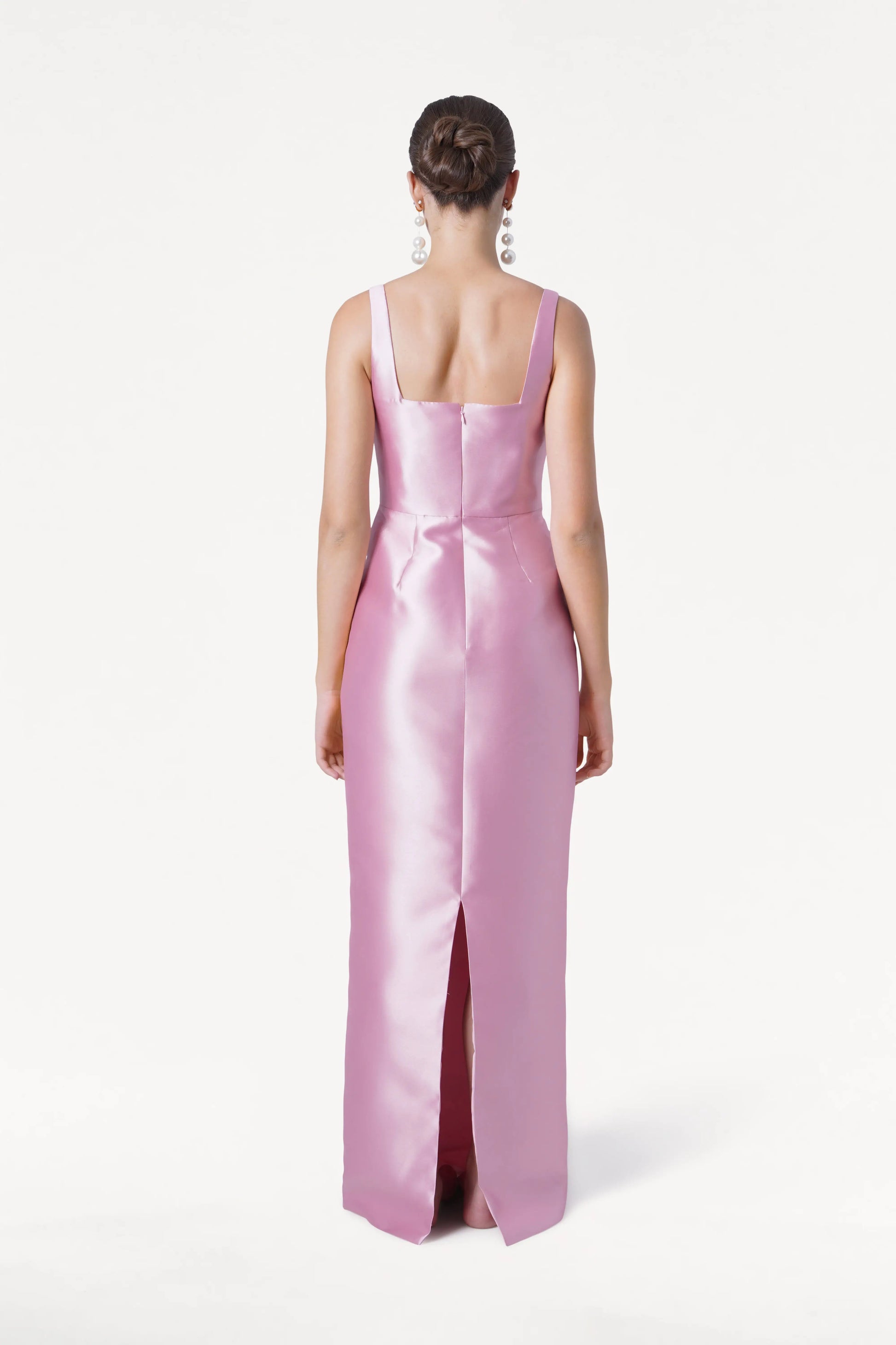 A woman with a neat bun and large dangling earrings faces away in the TH&TH Savoy Bridesmaid Dress in Bubblegum Pink Satin Twill, featuring wide straps, a center back seam, and a back slit against a plain white background.