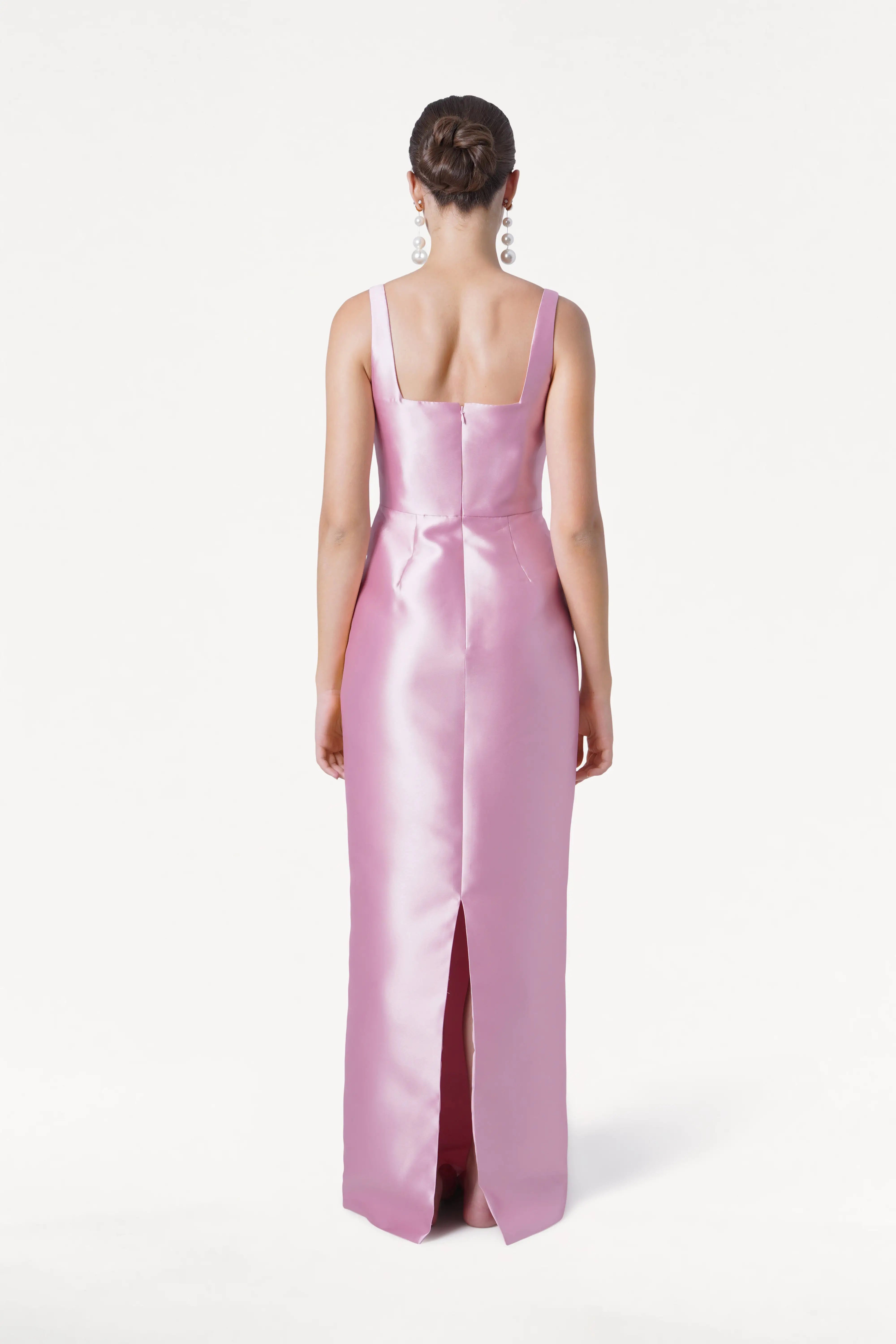 A woman with a neat bun and large dangling earrings faces away in the TH&TH Savoy Bridesmaid Dress in Bubblegum Pink Satin Twill, featuring wide straps, a center back seam, and a back slit against a plain white background.
