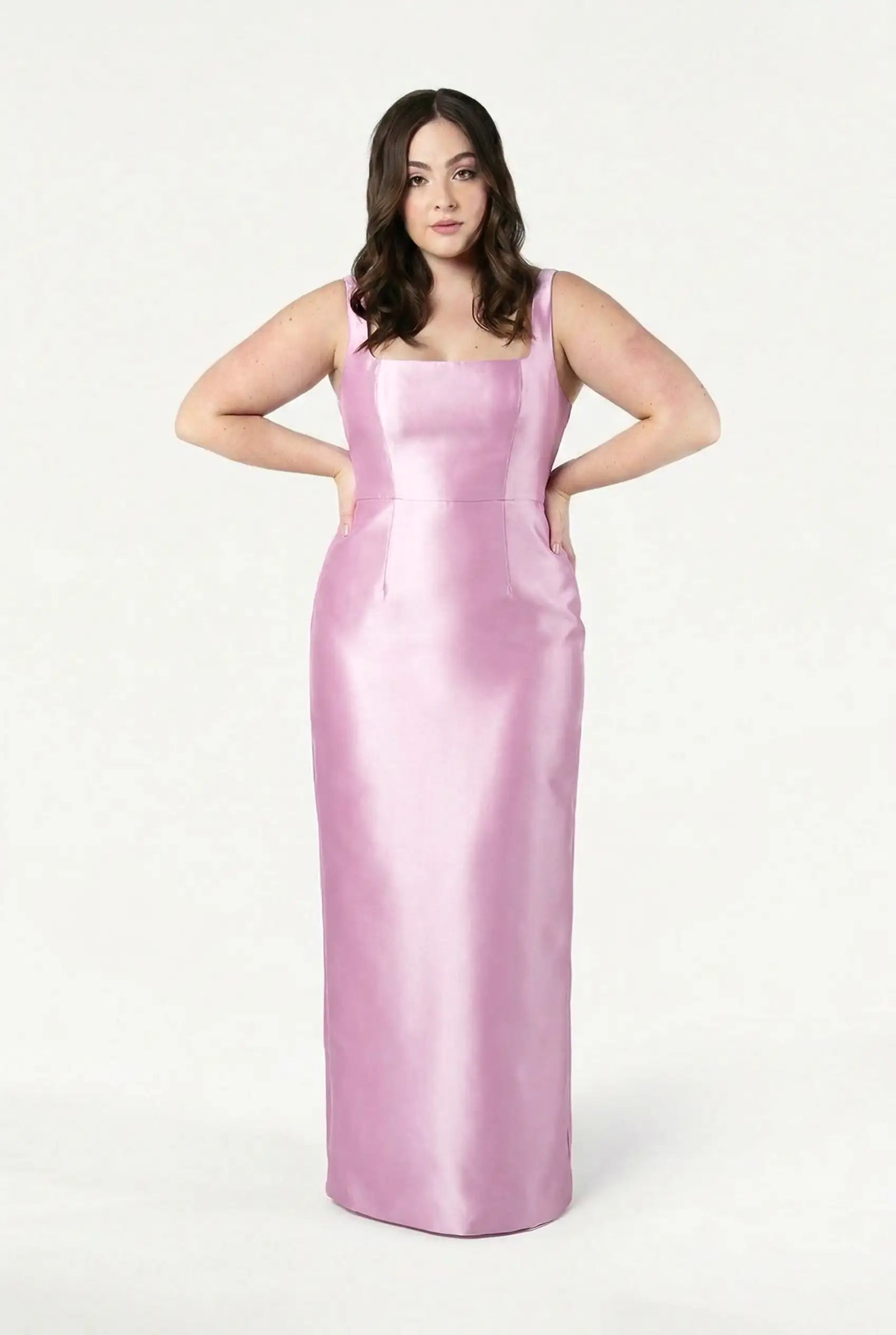 A woman with long brown hair poses against a white background, wearing the TH&TH Savoy Bridesmaid Dress in bubblegum pink satin twill, featuring a sleeveless, floor-length design and a square neckline.