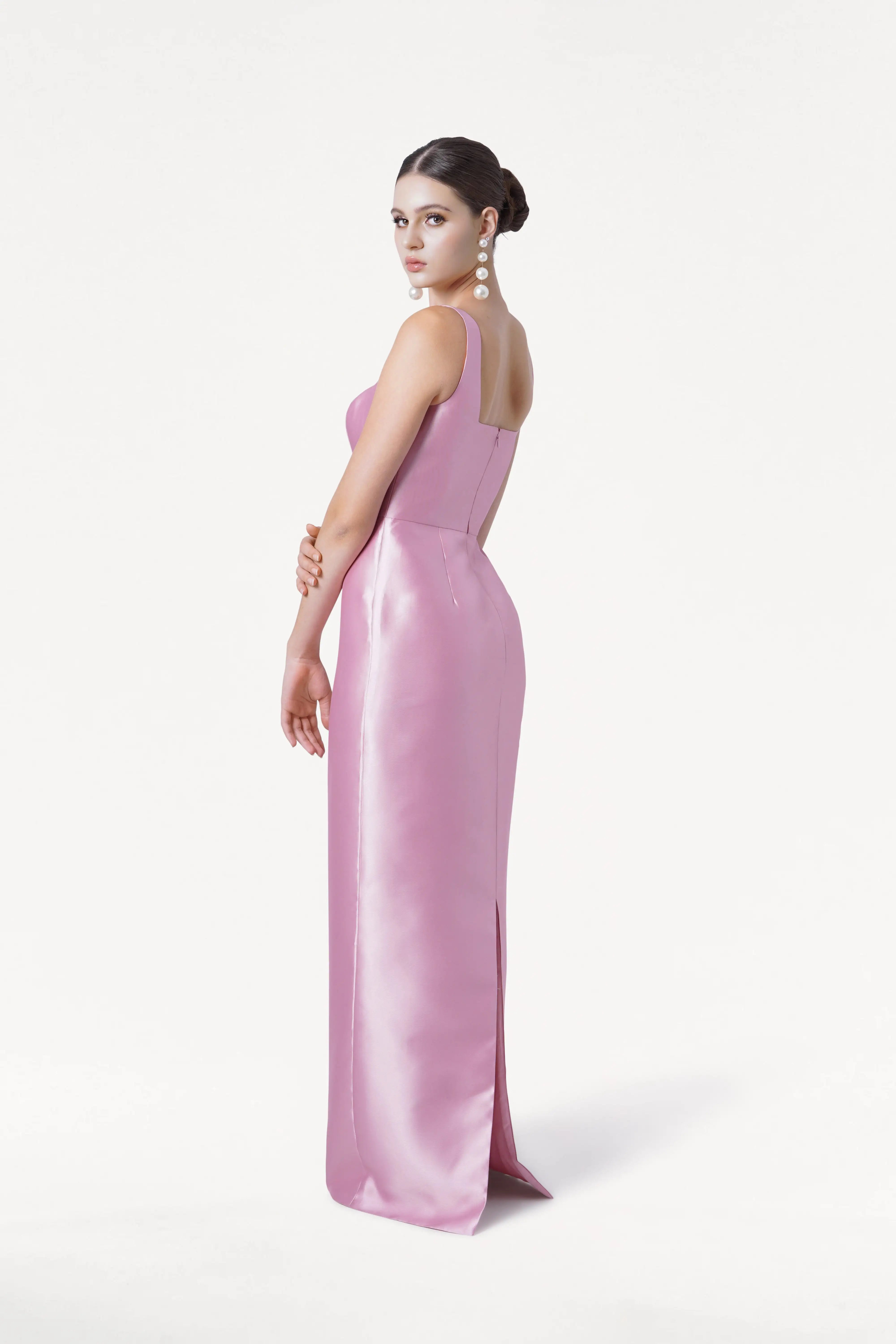 A woman in the TH&TH Savoy Bridesmaid Dress in Bubblegum Pink Satin Twill stands sideways against a white background, gazing at the camera. She accessorizes with large pearl earrings and her hair is styled in a neat bun.