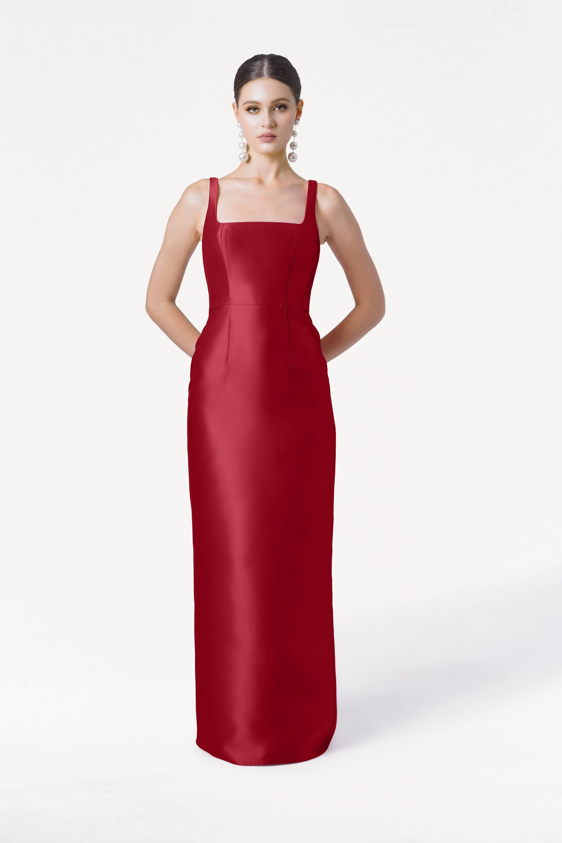 A woman stands against a white background in the TH&TH Savoy Bridesmaid Dress in Dark Cherry Satin Twill, a long, fitted, sleeveless gown with a modern square neckline. She wears large dangling earrings and has her hands behind her back.