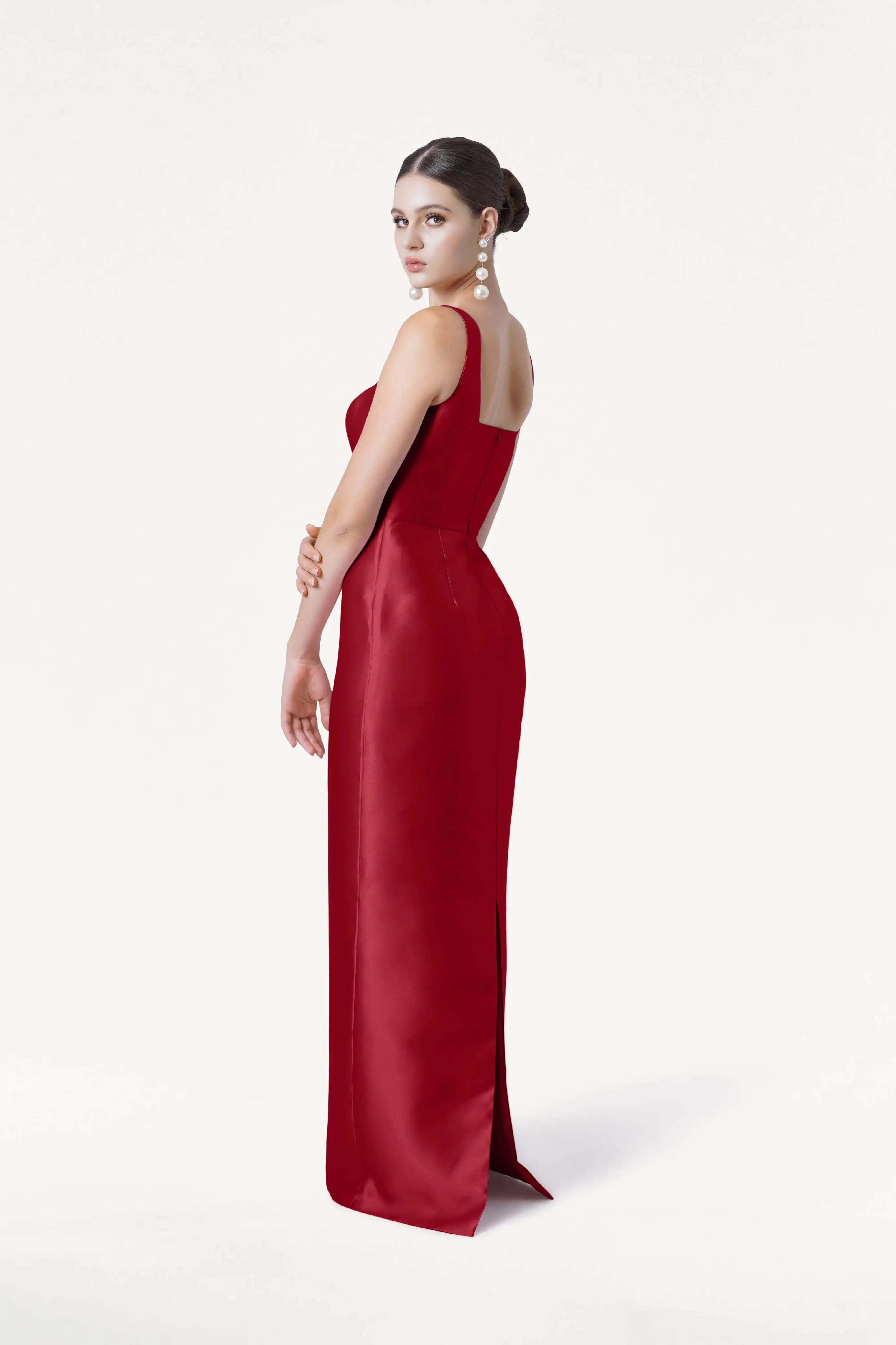 A woman models the TH&TH Savoy Bridesmaid Dress in Dark Cherry Satin Twill, a fitted floor-length gown. With her hair in a bun and wearing long earrings, she stands against a plain white background, glancing over her shoulder at the camera.