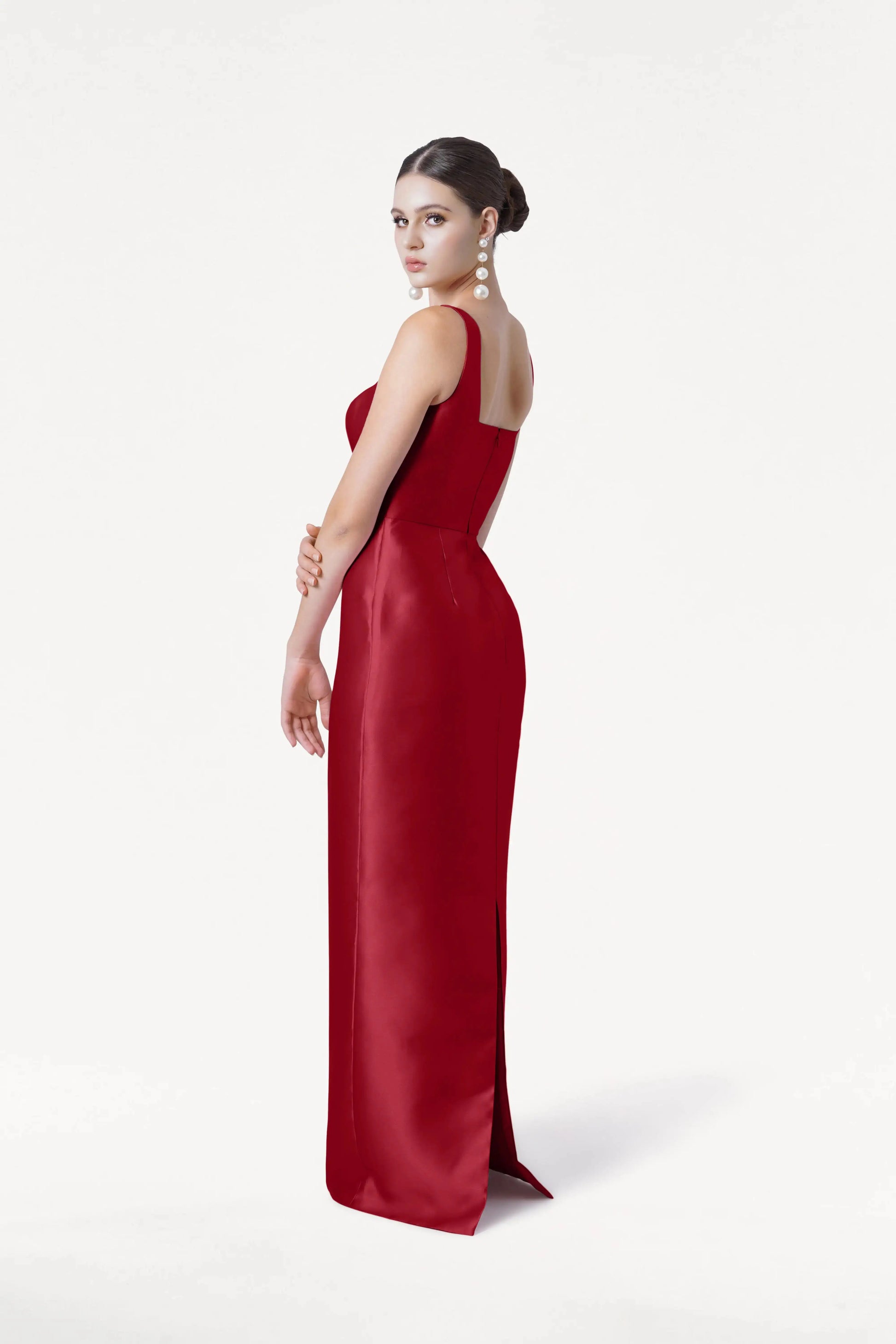 A woman models the TH&TH Savoy Bridesmaid Dress in Dark Cherry Satin Twill, a fitted floor-length gown. With her hair in a bun and wearing long earrings, she stands against a plain white background, glancing over her shoulder at the camera.