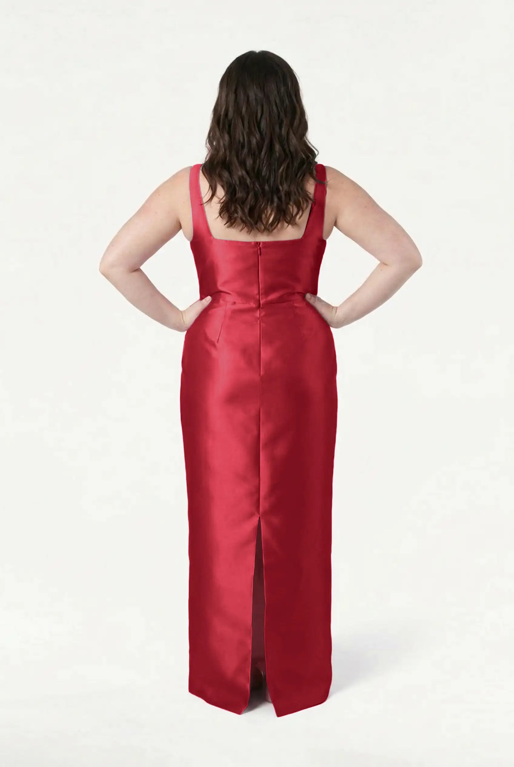 A woman with wavy brown hair, hands on hips, faces away in the TH&TH Savoy Bridesmaid Dress in Dark Cherry Satin Twill—a sleeveless, floor-length gown with a back slit and modern square neckline—set against a plain white background.