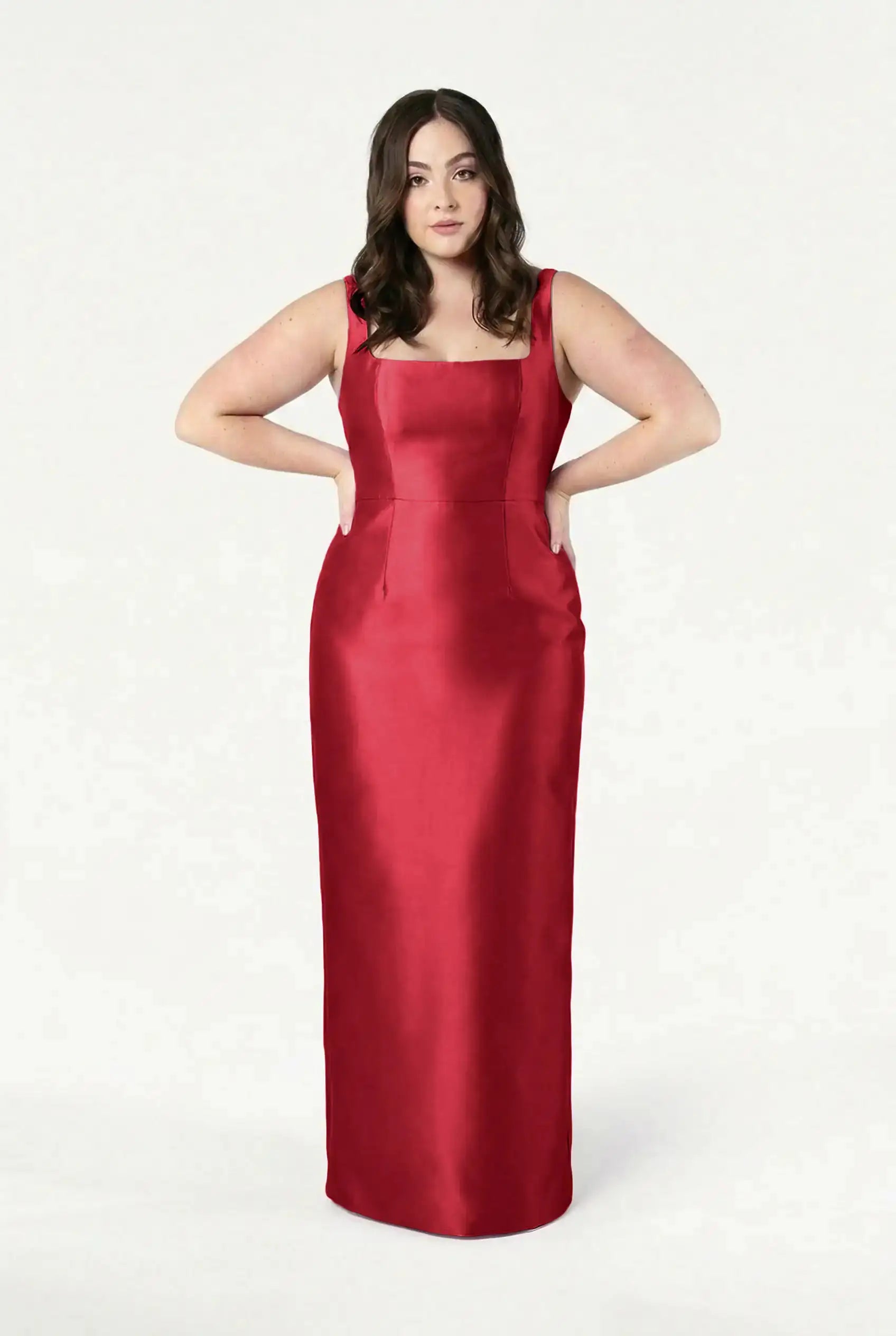 A woman poses against a white background, hands on hips, wearing the TH&TH Savoy Bridesmaid Dress in Dark Cherry Satin Twill—a sleeveless, floor-length gown with a modern square neckline.