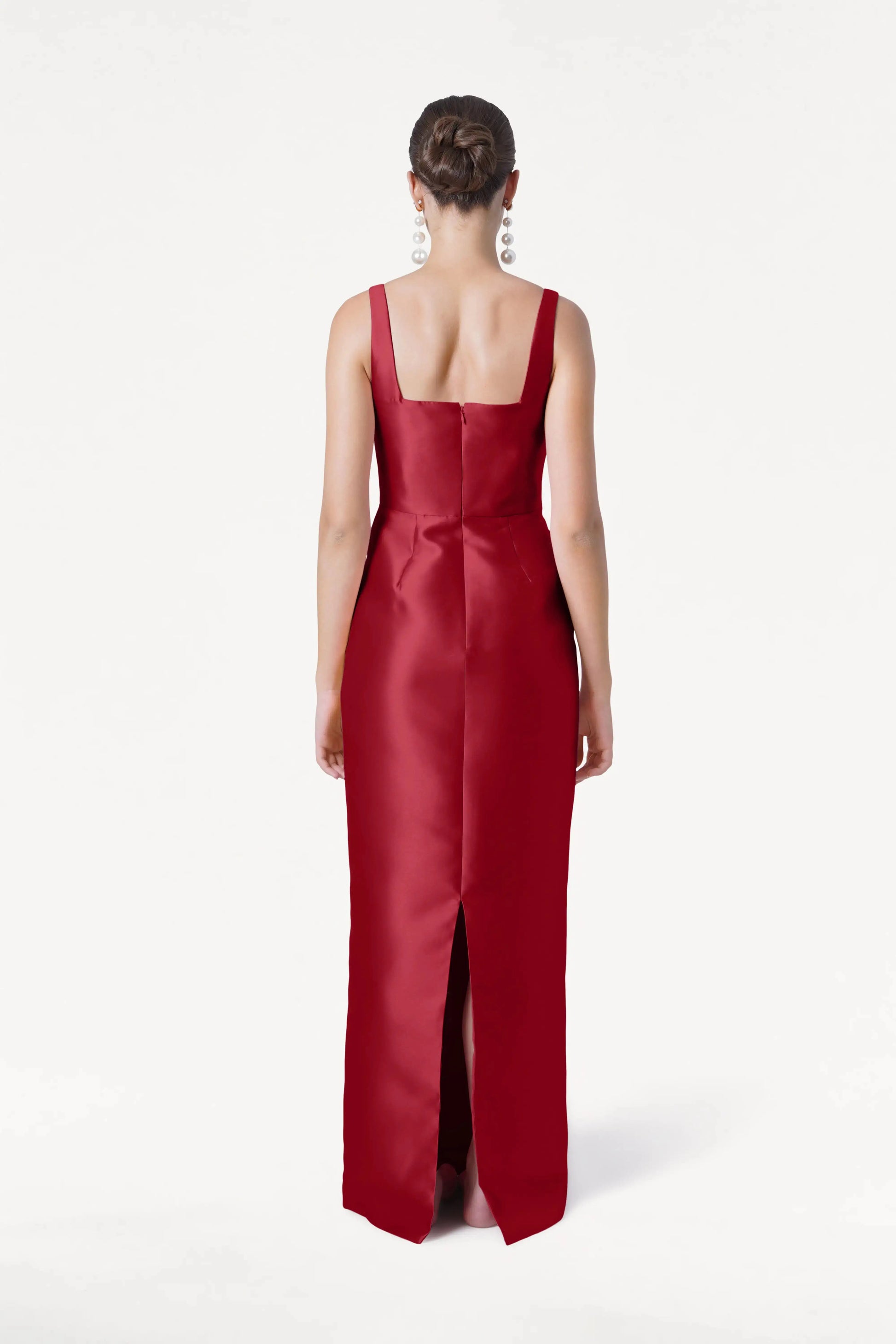 A woman with her hair in a bun models the TH&TH Savoy Bridesmaid Dress in Dark Cherry Satin Twill, featuring a sleeveless floor-length design with a square neckline and back slit, styled with statement earrings, shown from the back against a white backdrop.