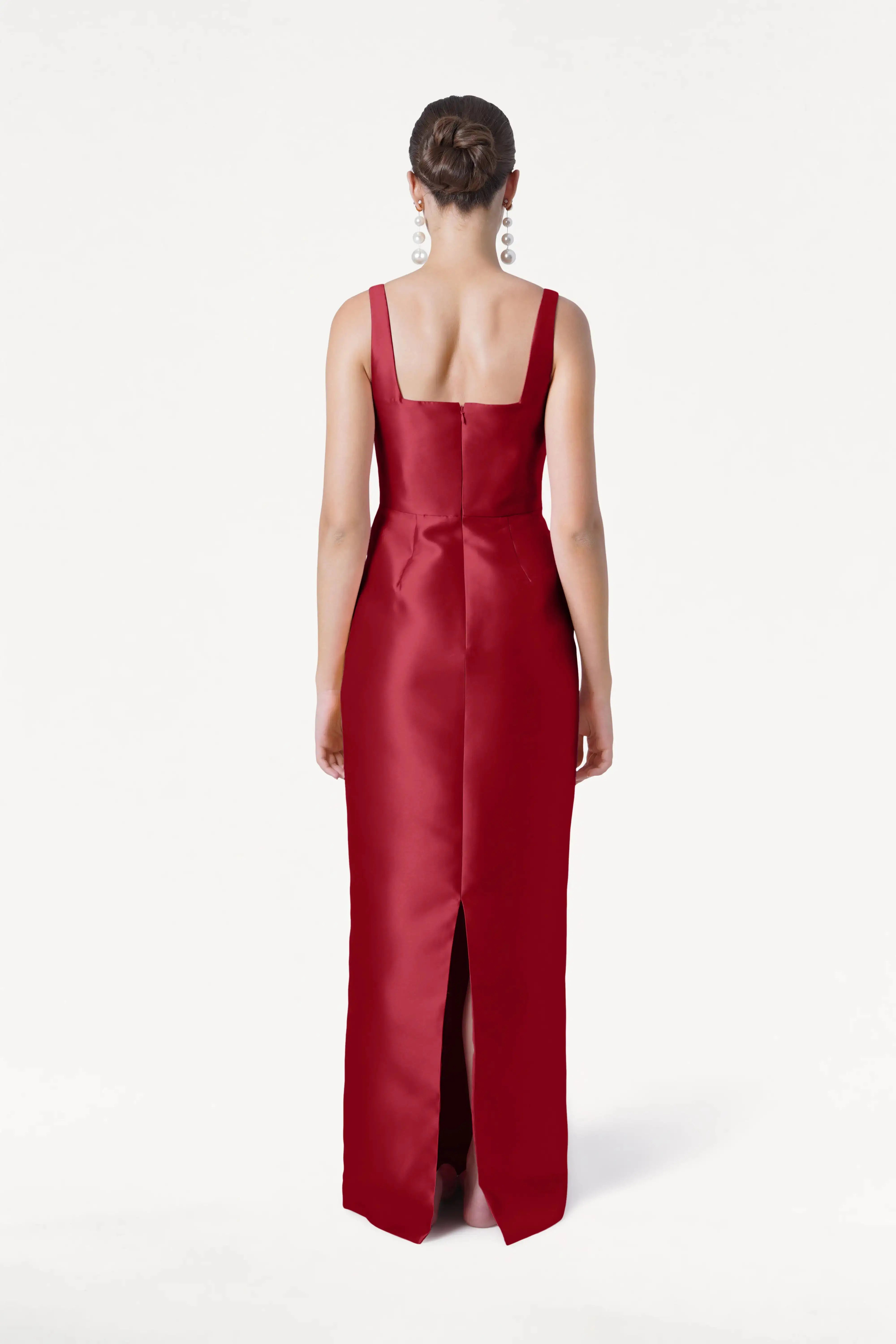 A woman with her hair in a bun models the TH&TH Savoy Bridesmaid Dress in Dark Cherry Satin Twill, featuring a sleeveless floor-length design with a square neckline and back slit, styled with statement earrings, shown from the back against a white backdrop.