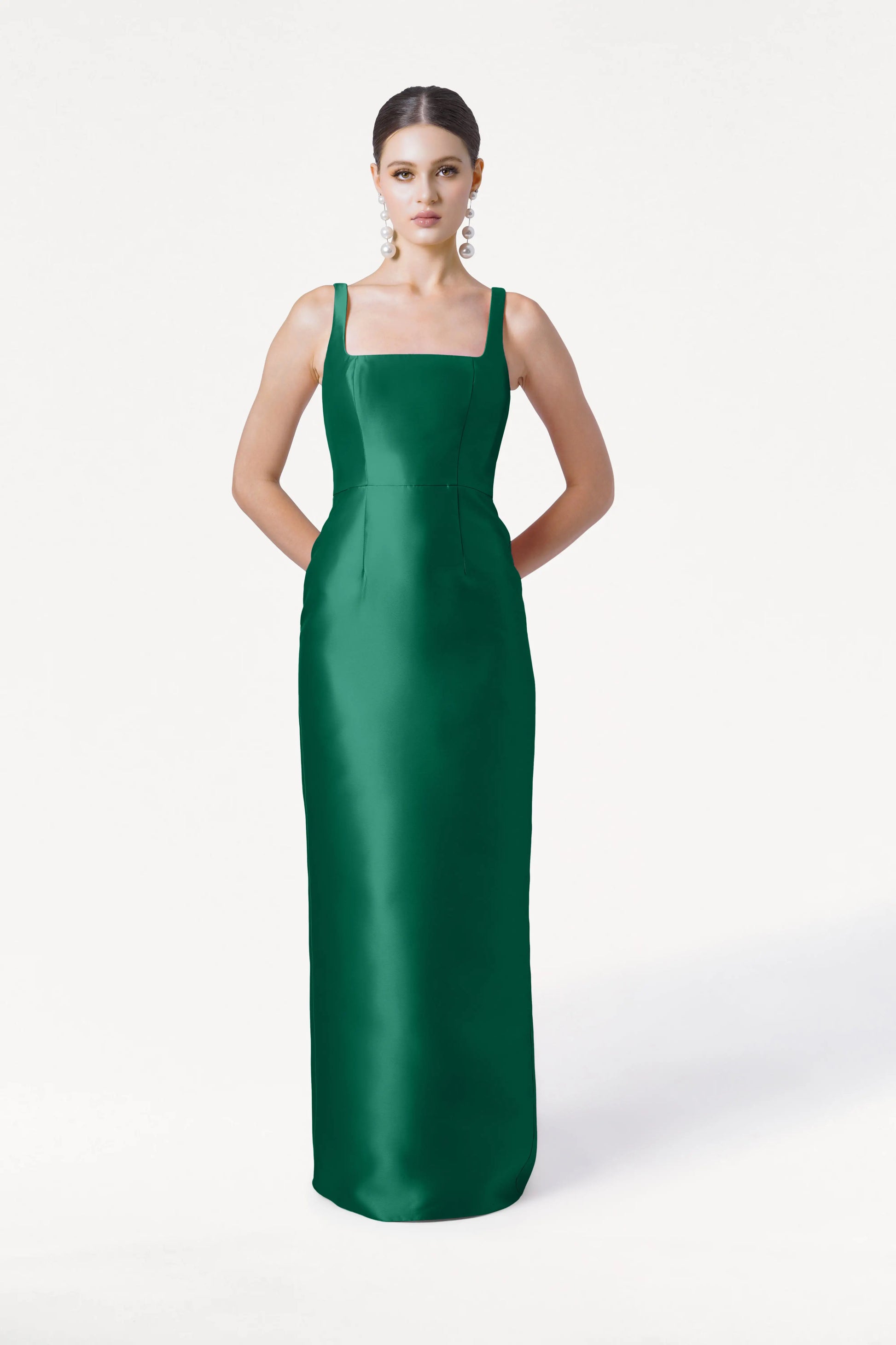 A woman poses against a white background in the TH&TH Savoy Bridesmaid Dress in Forest Green Satin Twill, featuring a sleeveless, floor-length design and square neckline. She wears statement earrings with her hands behind her back.