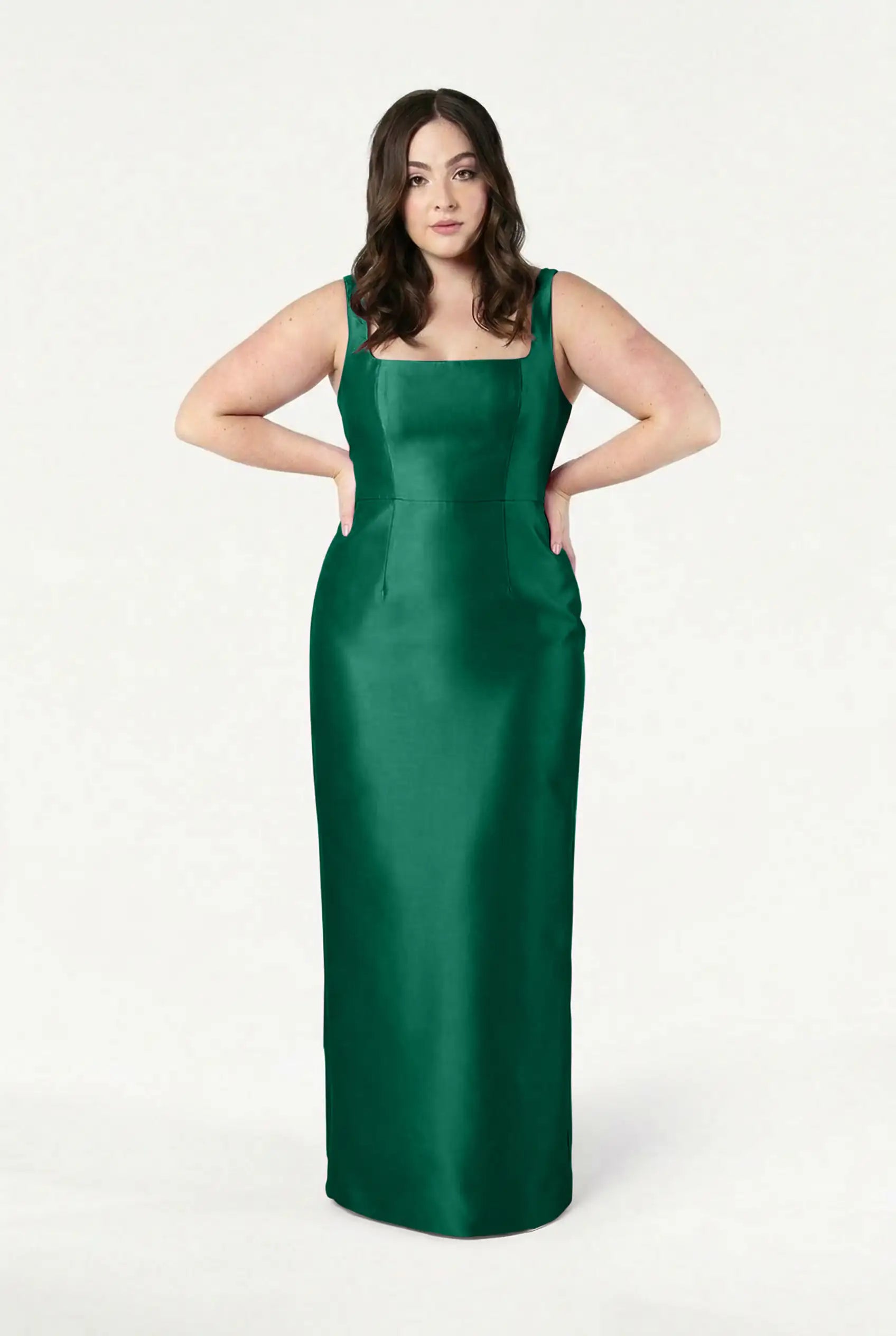 A woman with long brown hair stands against a white background, wearing the TH&TH Savoy Bridesmaid Dress in Forest Green Satin Twill—a sleeveless, floor-length gown with a square neckline and her hands on her hips.