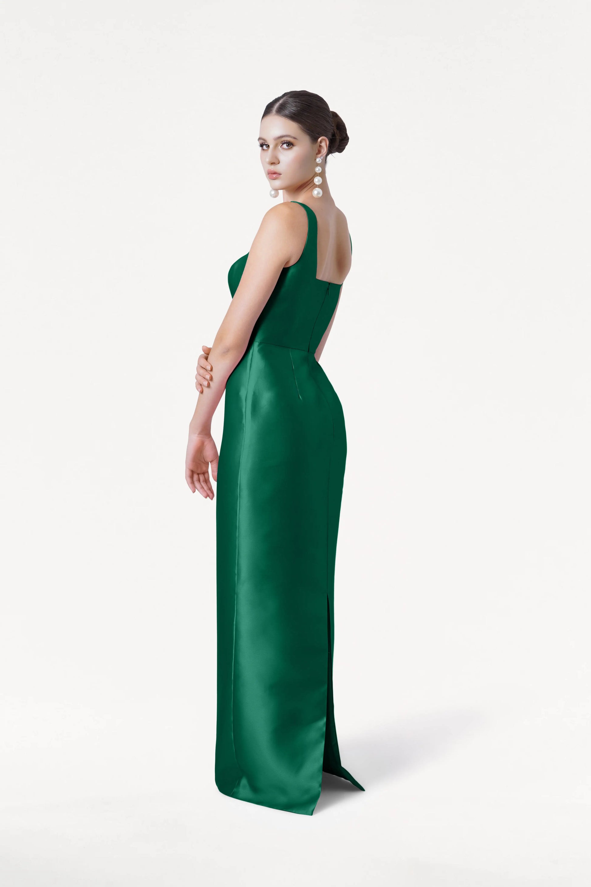 A woman in the TH&TH Savoy Bridesmaid Dress in Forest Green Satin Twill stands against a white backdrop, looking over her shoulder with her hair in a sleek bun and statement drop earrings.