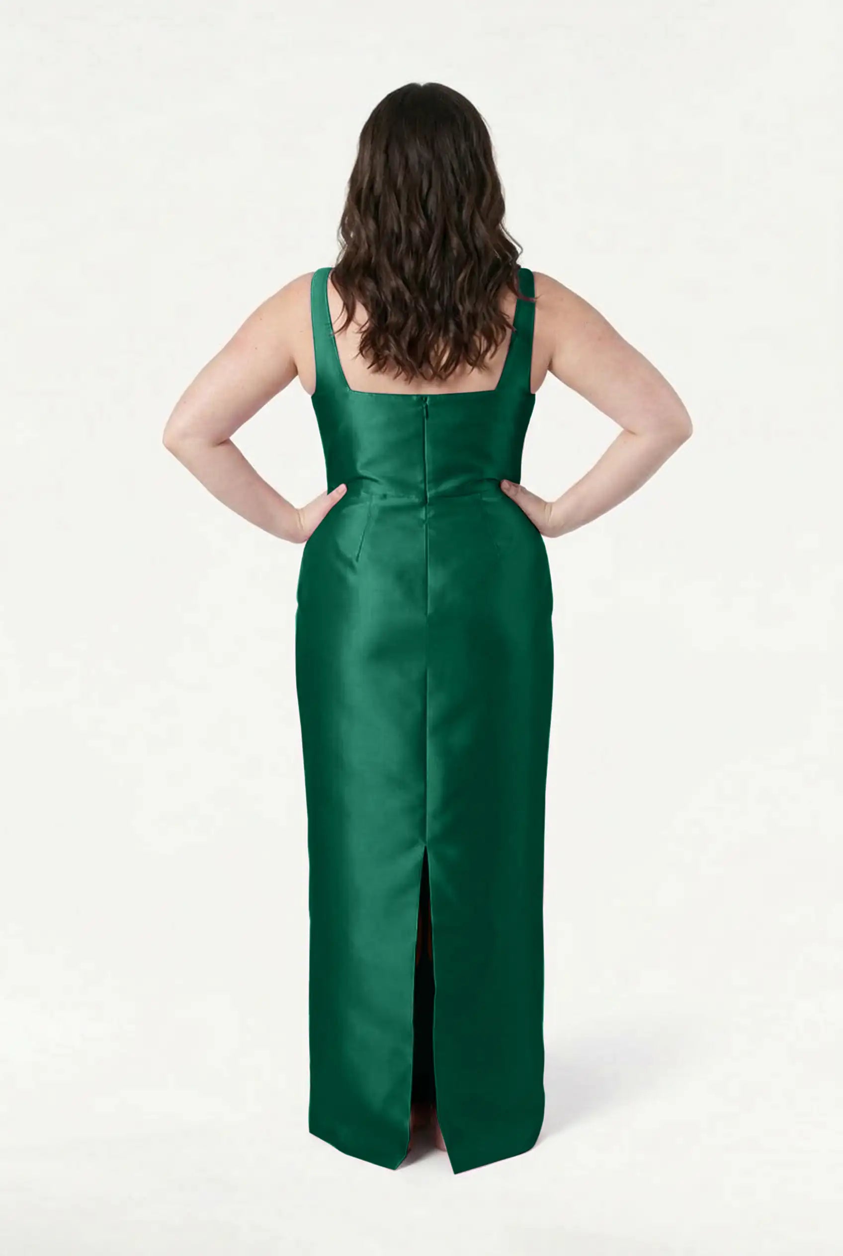A woman faces away with hands on hips, wearing the TH&TH Savoy Bridesmaid Dress in Forest Green Satin Twill—a sleeveless, floor-length gown with a back slit. Her dark wavy hair falls to her shoulders against a plain white background.
