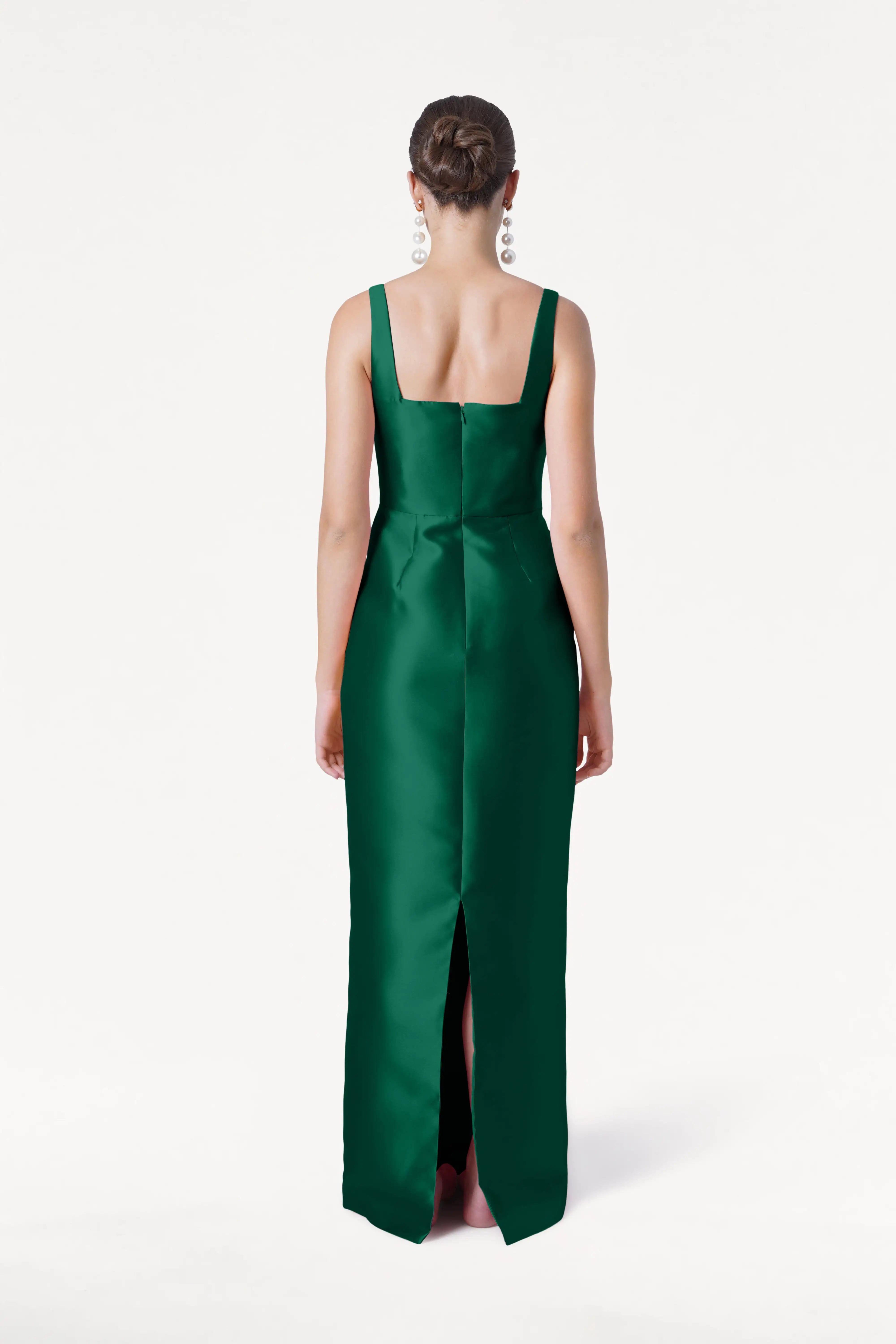 A woman with her hair in a bun faces away, wearing the TH&TH Savoy Bridesmaid Dress in Forest Green Satin Twill—a sleeveless, floor-length gown with a back slit—set against a plain white background.
