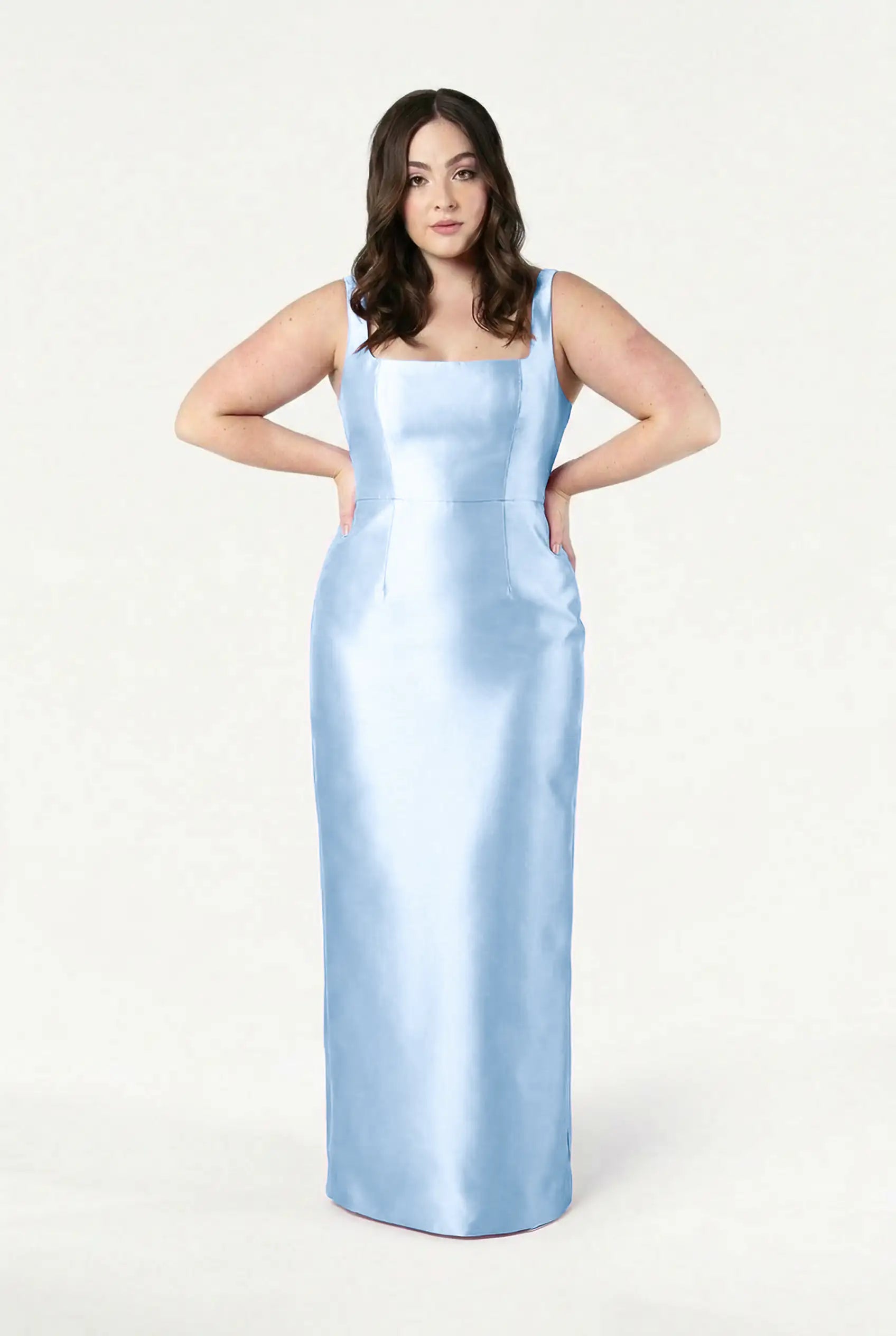 A woman with long brown hair models the TH&TH Savoy Bridesmaid Dress in frosted blue satin twill, featuring a sleeveless square neckline, as she stands confidently with her hands on her hips against a plain white background.