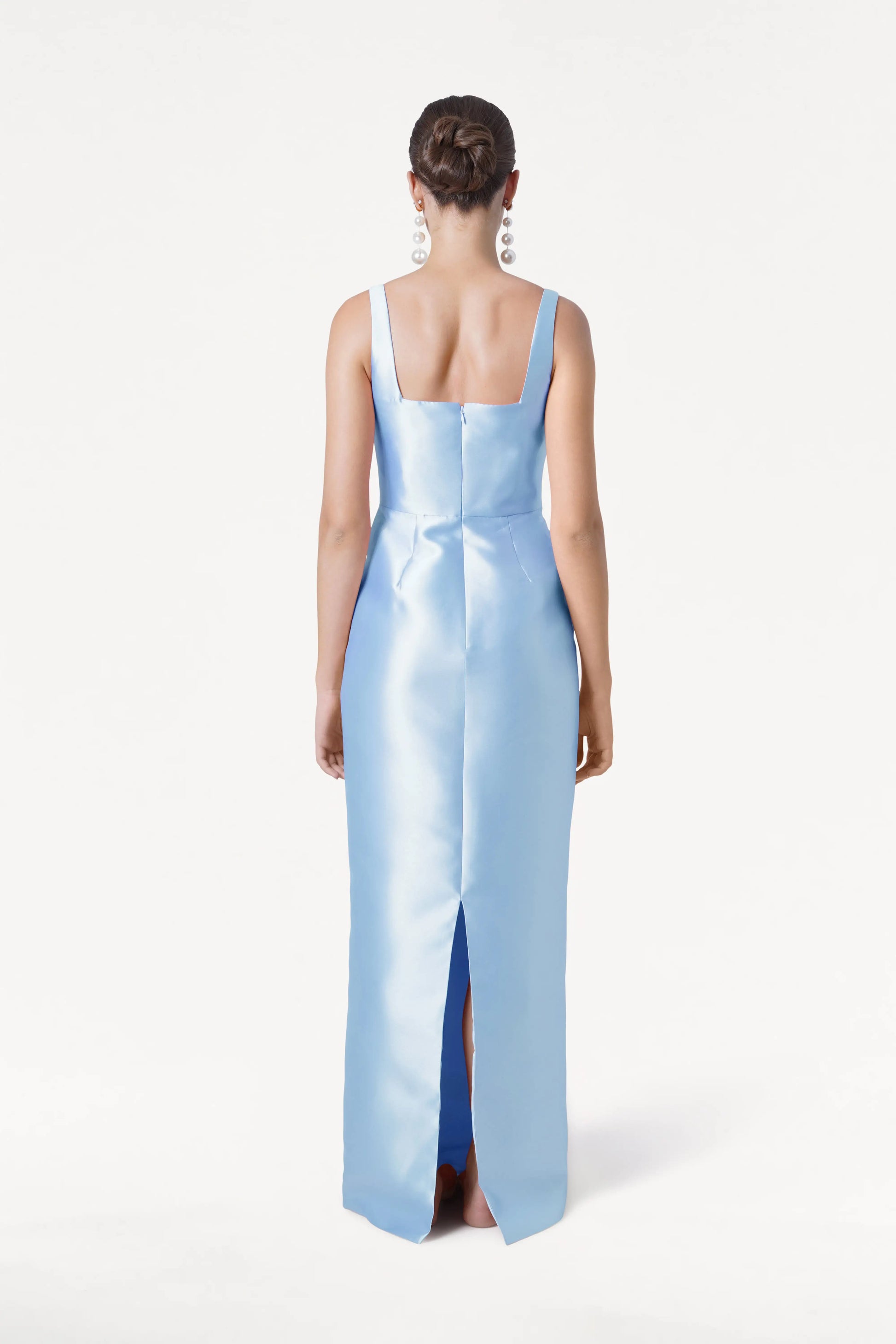 A woman with a neat bun and large earrings faces away in the TH&TH Savoy Bridesmaid Dress, crafted from frosted blue satin twill. The gown features a square neckline, thick straps, and a back hem slit.