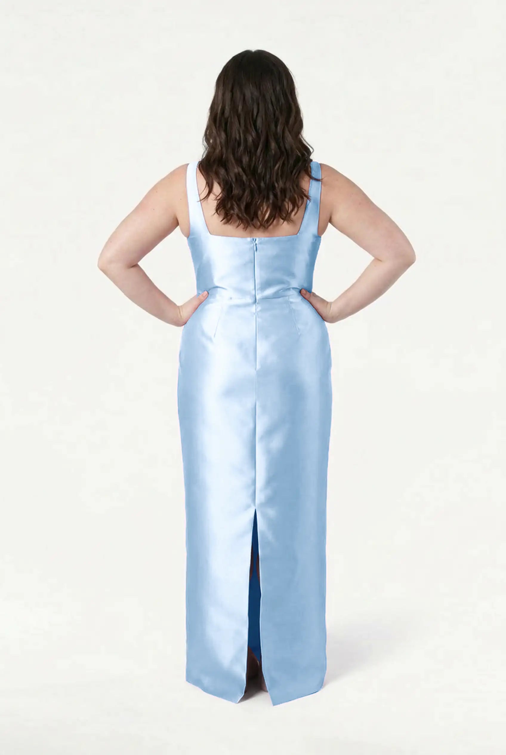 A woman with wavy brown hair, facing away, wears the TH&TH Savoy Bridesmaid Dress in Frosted Blue Satin Twill—a sleeveless, floor-length gown with a back slit—set against a plain white background.