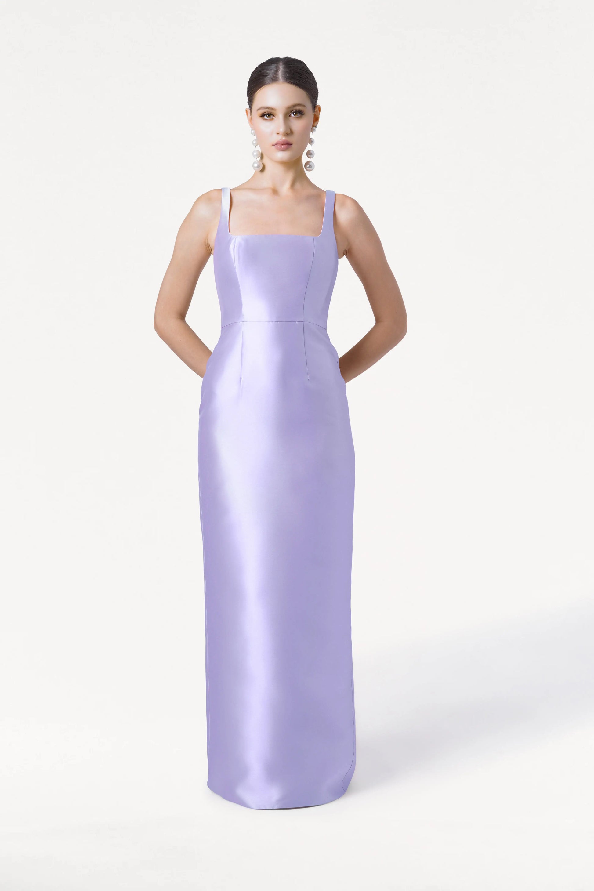 A woman faces forward in the Savoy Bridesmaid Dress by TH&TH—a sleeveless, floor-length lilac mist satin twill gown with thin straps. Hands behind her back, she wears pearl drop earrings against a plain white background.