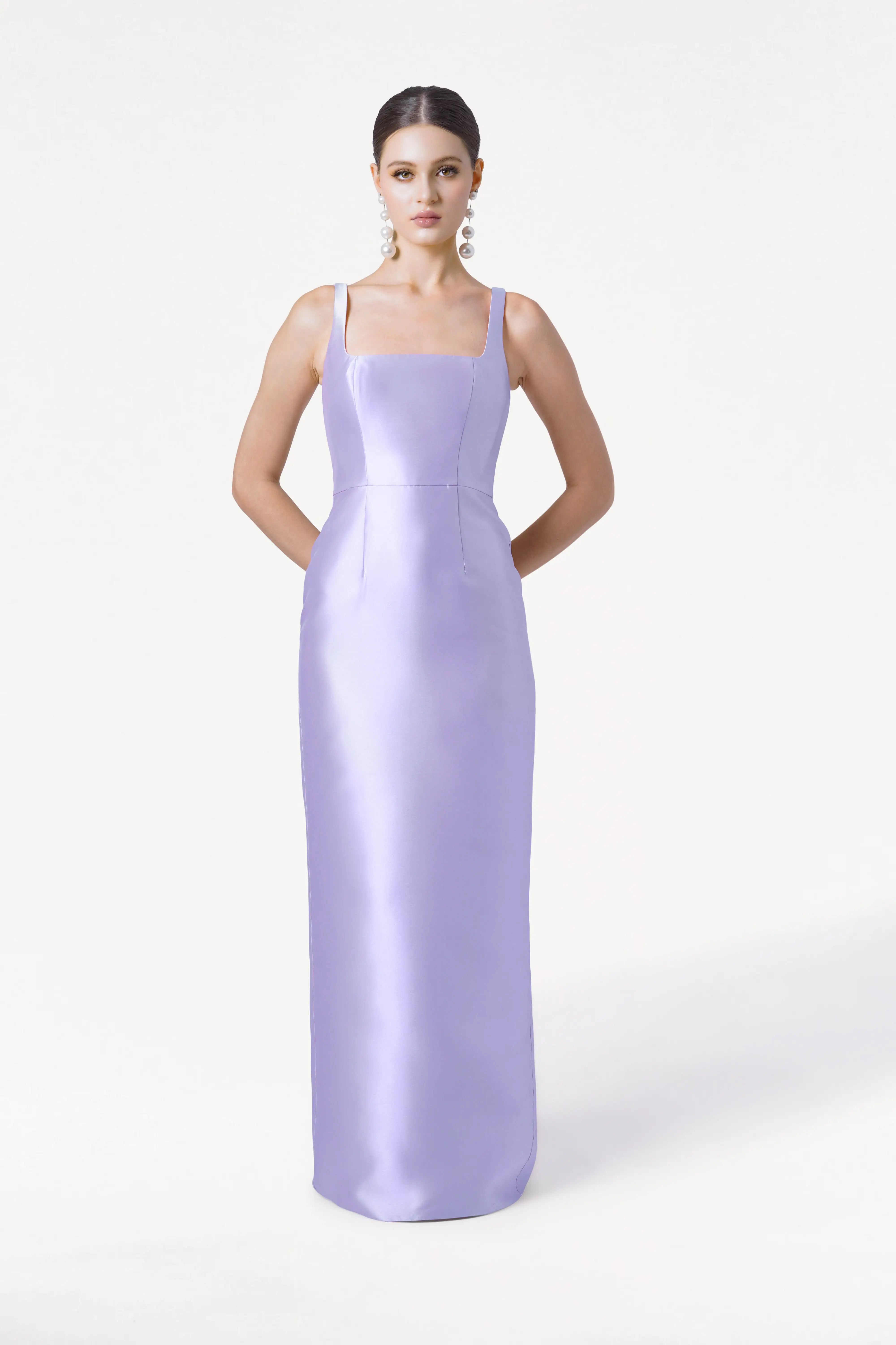 A woman faces forward in the Savoy Bridesmaid Dress by TH&TH—a sleeveless, floor-length lilac mist satin twill gown with thin straps. Hands behind her back, she wears pearl drop earrings against a plain white background.
