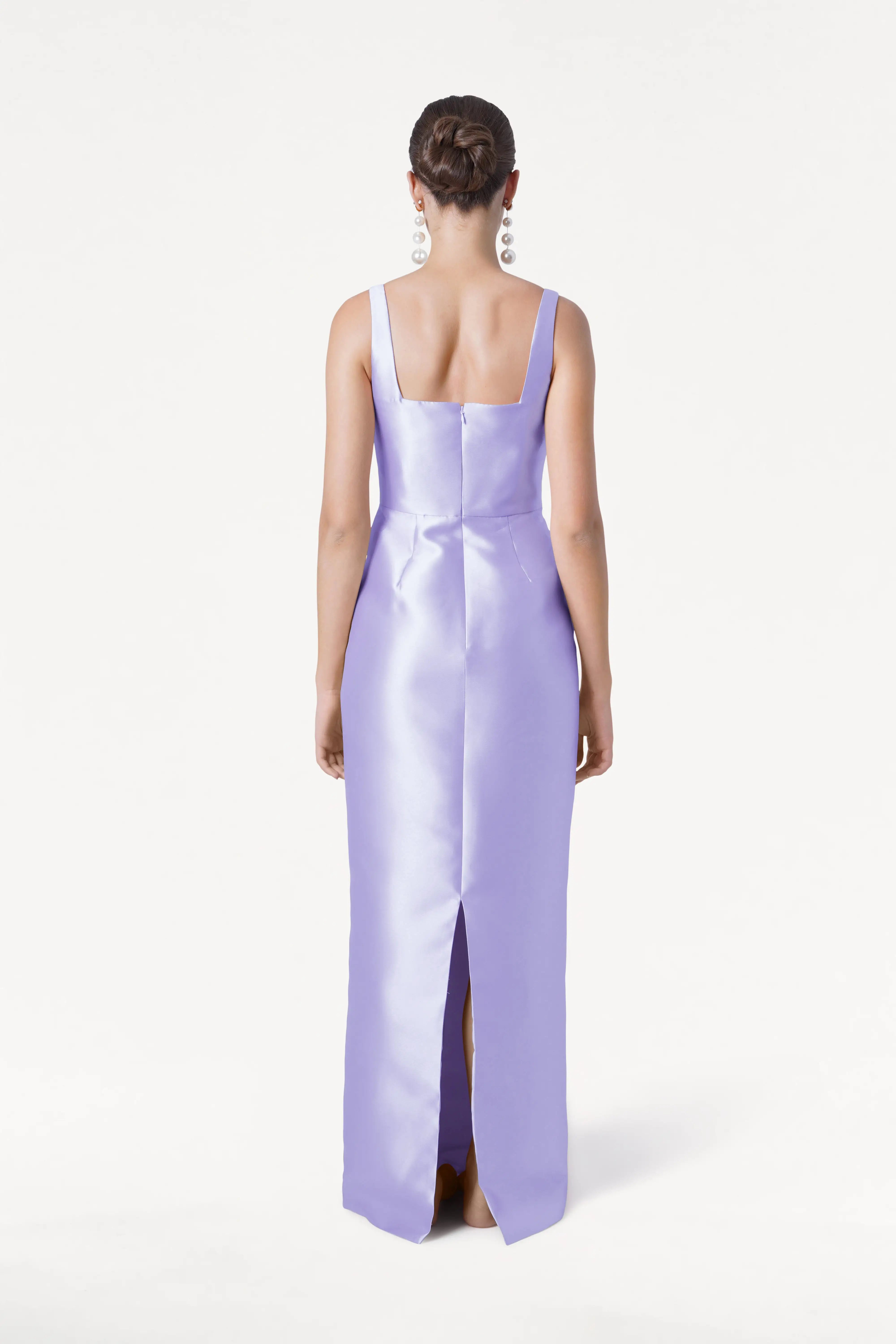 A woman with a bun and dangling earrings stands against a white background, wearing the TH&TH Savoy Bridesmaid Dress in Lilac Mist Satin Twill—a fitted, floor-length, sleeveless gown with a square neckline and back slit.